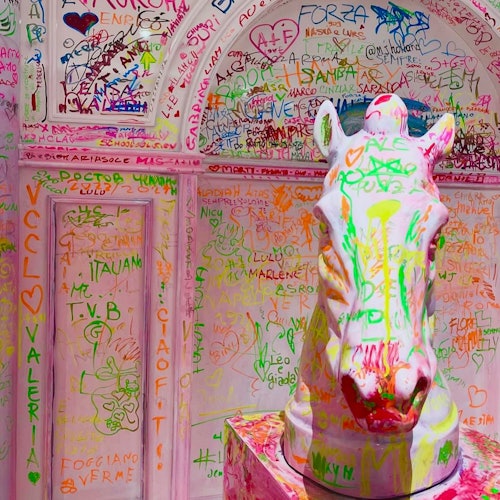 Una statua di cavallo bianco è coperta da graffiti colorati, circondata da un muro con vari nomi, frasi e disegni in colori vivaci.