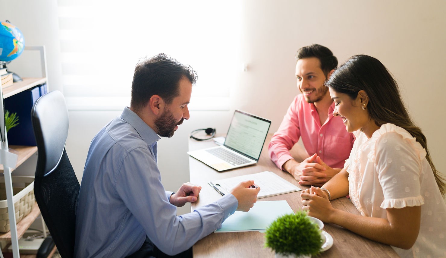 A couple consults with a professional advisor in a bright, welcoming office. They are engaged and reviewing documents together on a laptop.
