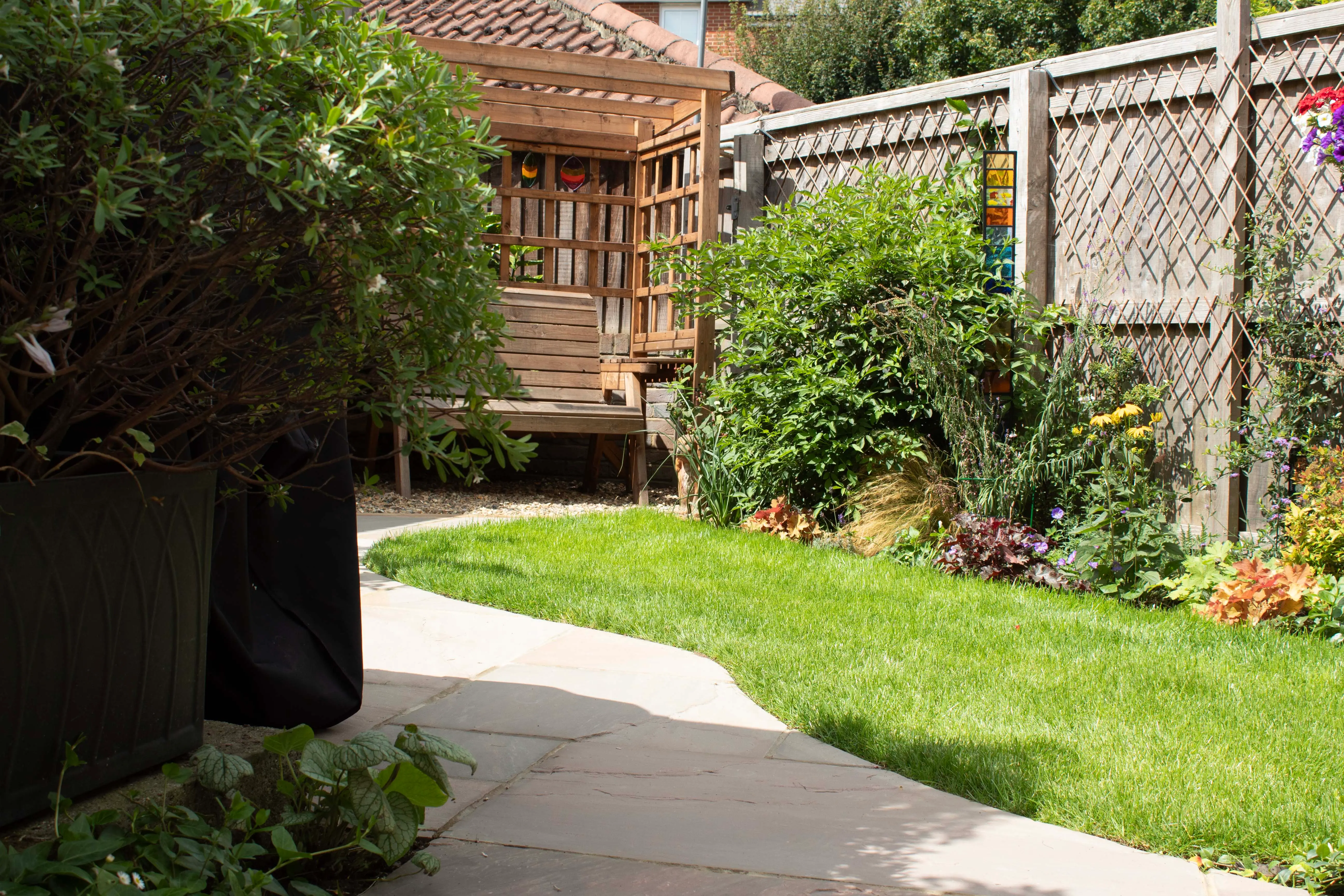 A grassy pathway leads to a wooden structure surrounded by greenery and garden plants in a sunny setting.