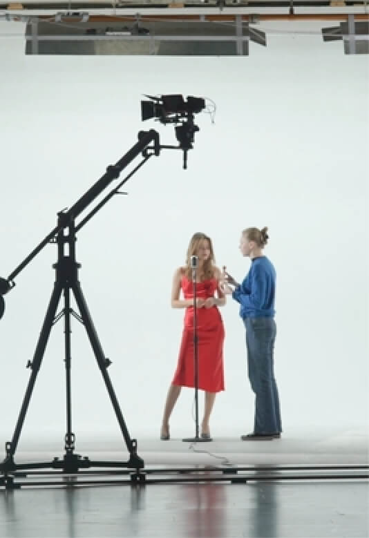 A woman in a red dress and a man in a blue shirt interact in a studio with a tripod camera.