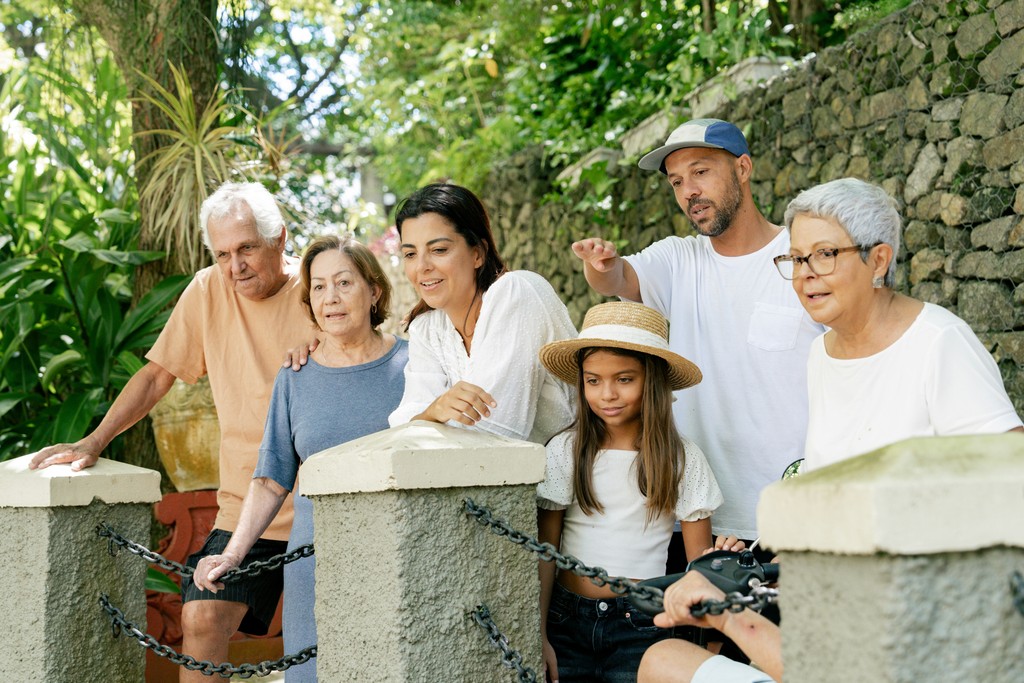  A multi-generational family gathered outdoors by a stone wall, engaged in a shared activity, with grandparents, parents, and children enjoying a sunny day together amidst lush greenery.