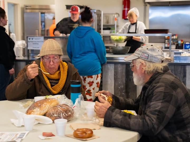 Two older men sit at a table in a community kitchen, bundled in warm clothes and sharing a meal with bread, salad, and hot food on disposable plates. Behind them, volunteers serve food from a counter while others wait in line, creating a welcoming and supportive atmosphere.