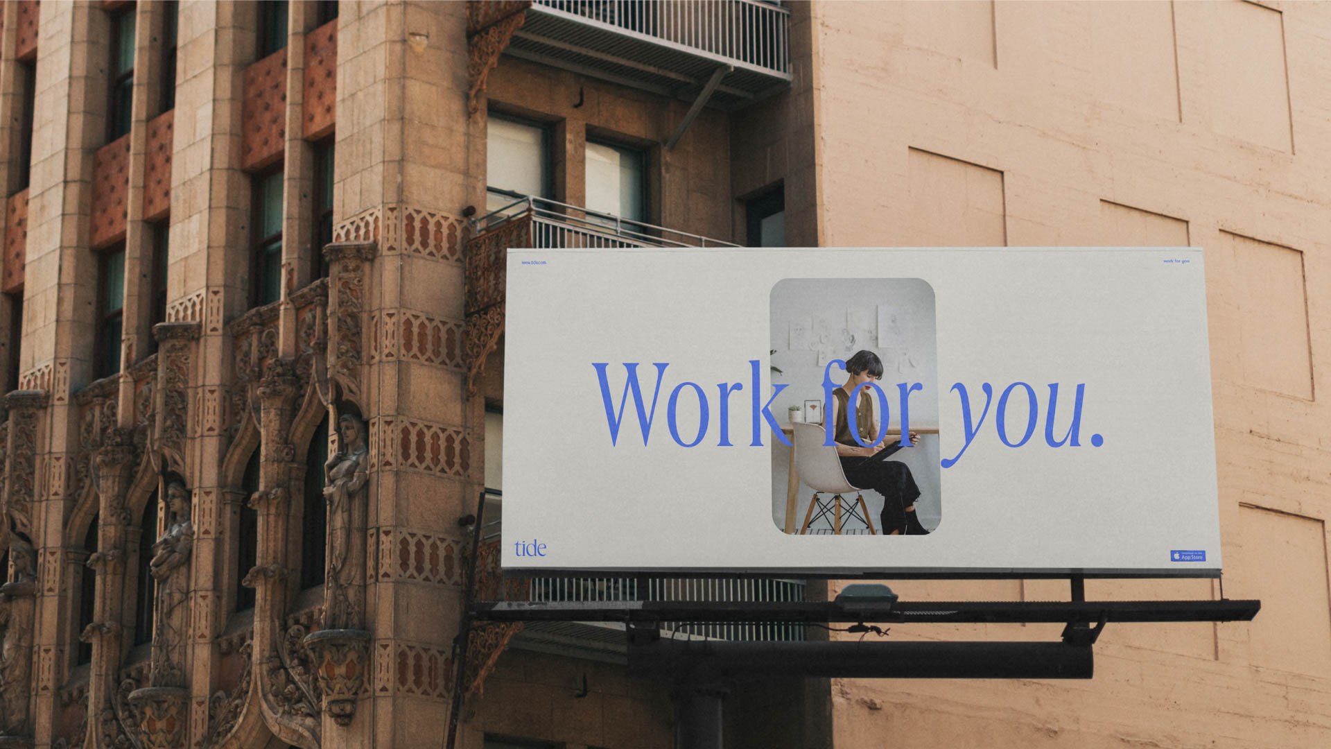 The image shows a billboard mounted on the side of a light brown brick building. The billboard is white with blue text that reads "Work for you." A smaller Tide logo is visible in the bottom right corner. Centered on the billboard is an illustration of a person sitting at a desk, working on a laptop. The building features decorative stonework on its facade.