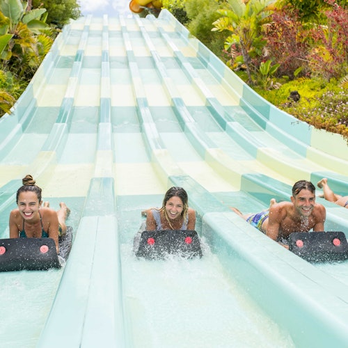 Three people slide down parallel water slides on mats, smiling and surrounded by lush greenery.