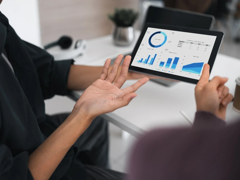 Two people in a meeting reviewing business analytics charts on a tablet, including bar graphs and a donut chart