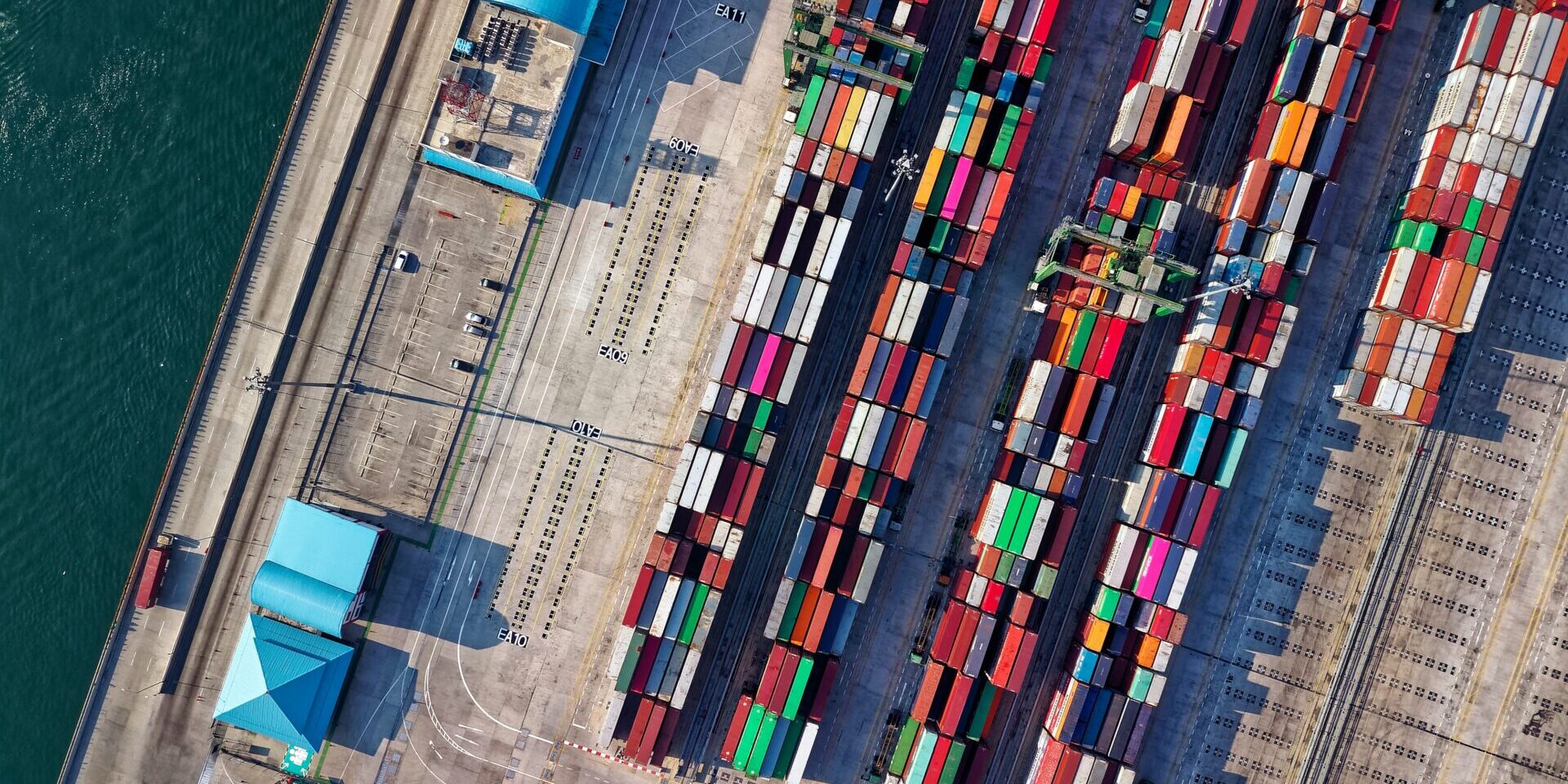 Aerial view of a busy shipping port featuring a variety of colorful shipping containers arranged in neat rows beside turquoise waters.