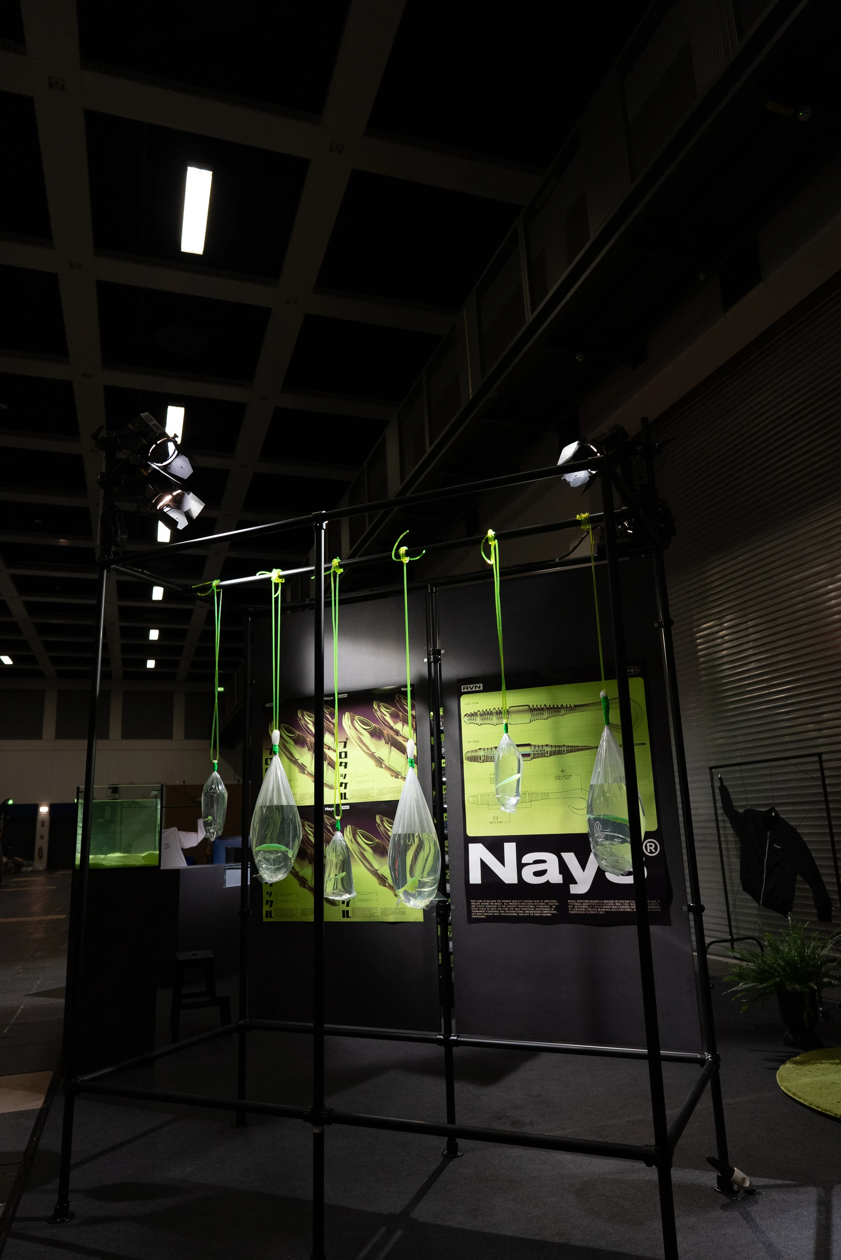 Black scaffold frame of the Nays booth with hanging water bags, green straps and backlit brand posters in the dark hall.