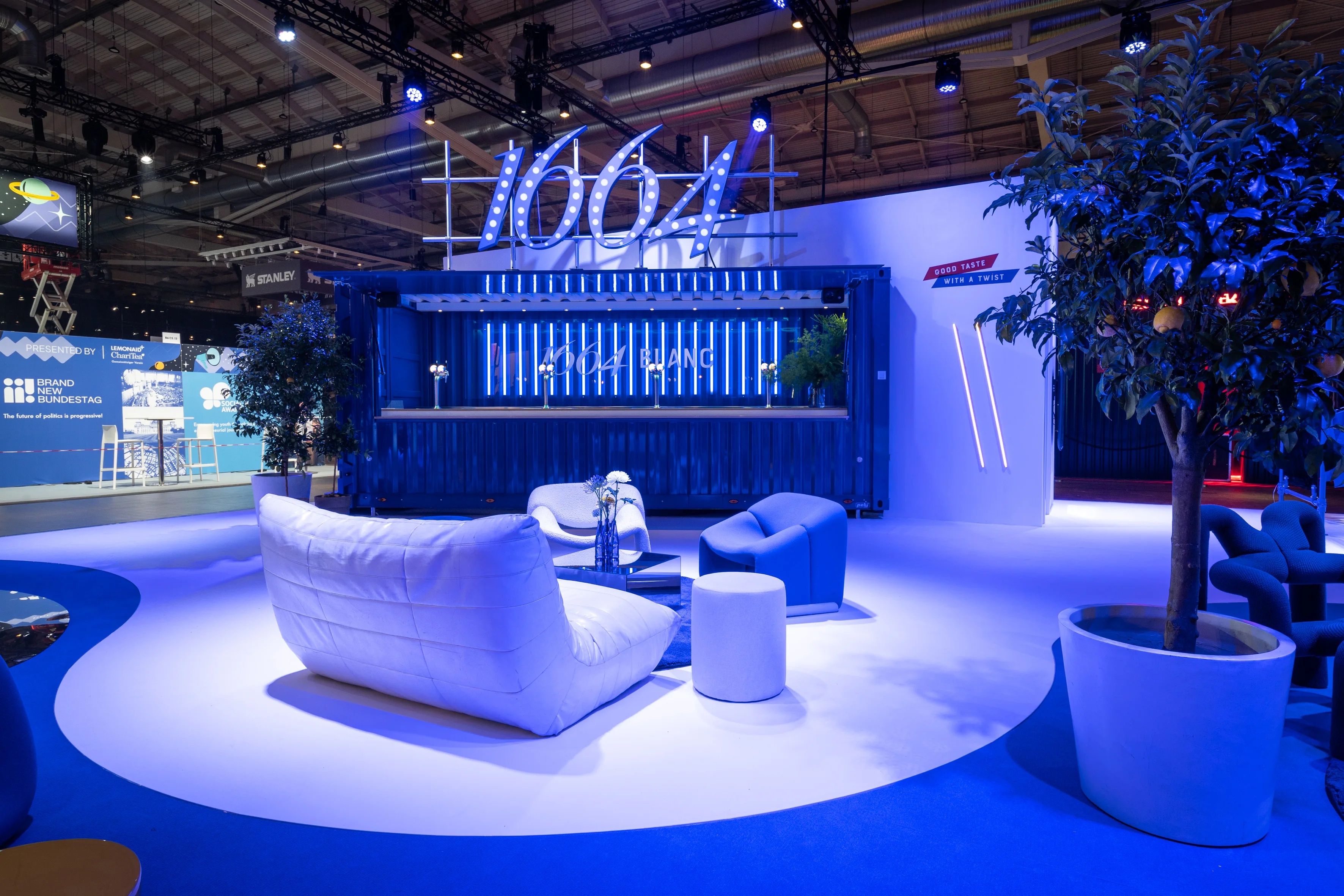 White lounge sofa and armchairs in front of the blue shipping-container bar with glowing 1664 logo at OMR.