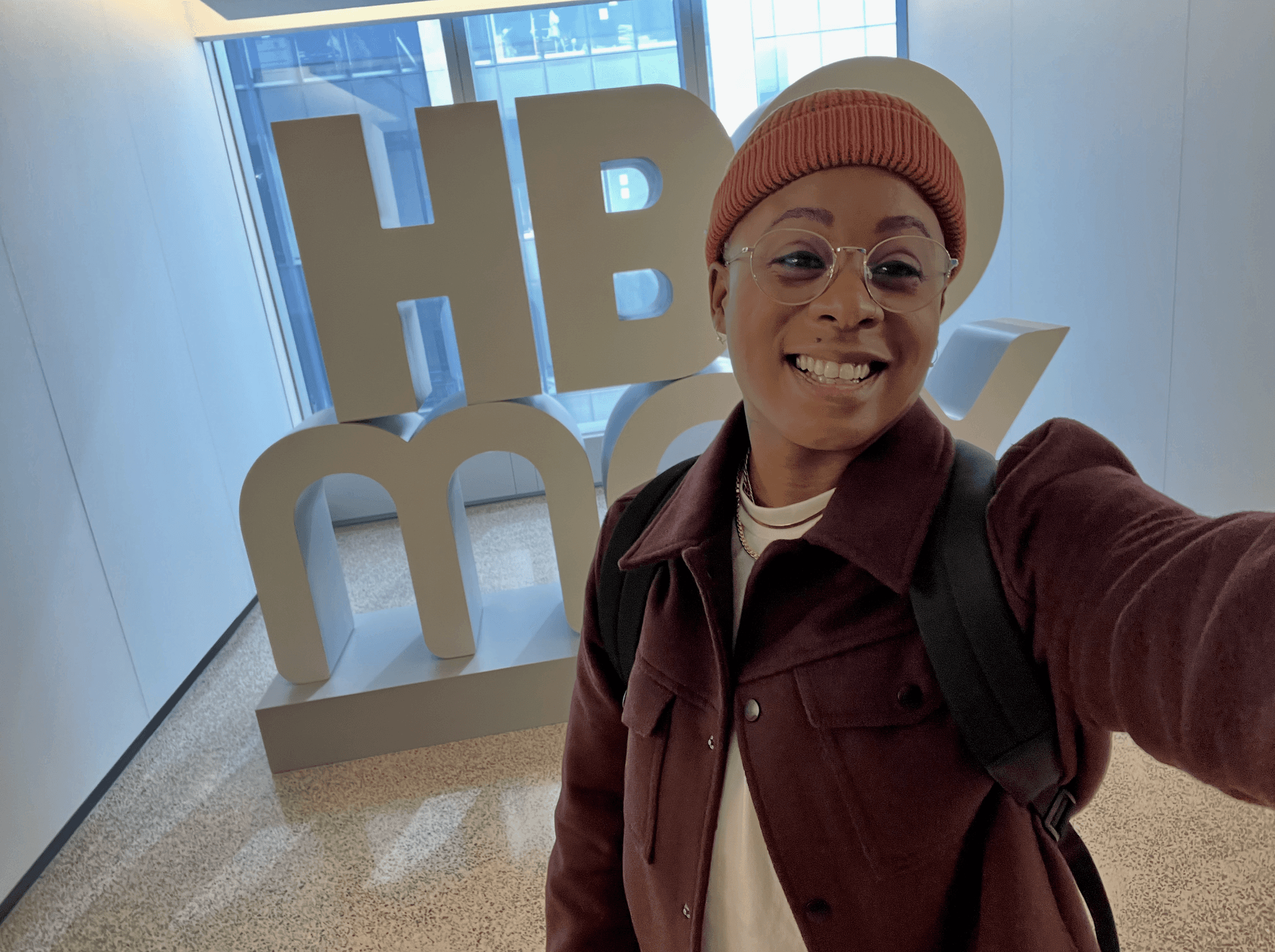 Ashleigh Warren smiles for a selfie in front of a large "HBOMax" sign in a well-lit room.