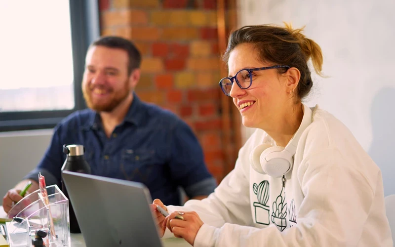 A photo of a woman wearing glasses with her brown hair tied up wearing a white jumper sits in front of a laptop and is smiling at something out of frame, in the background a man in a blue shirt is also smiling at the same focus point