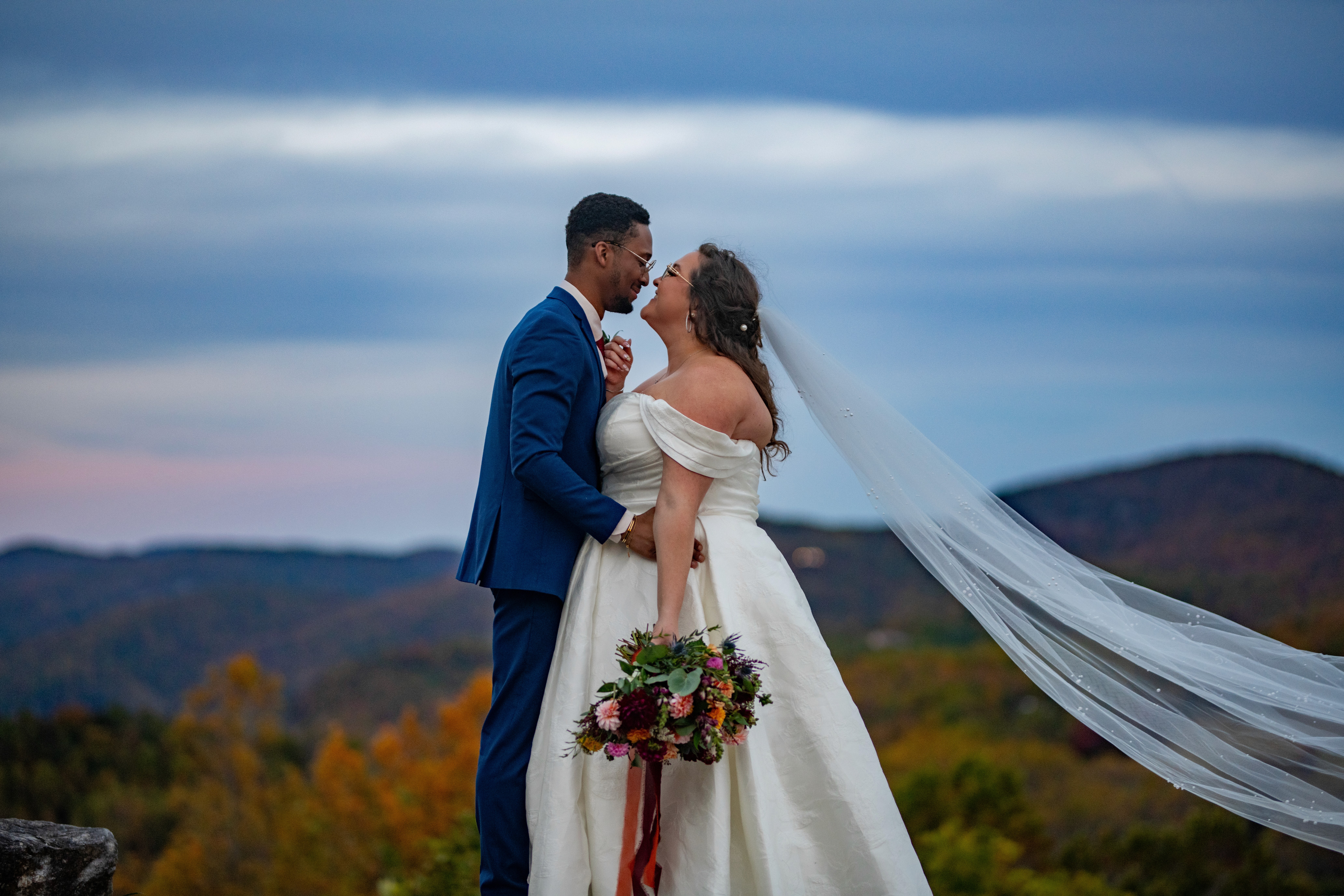 Couple dancing outdoors during golden hour. Woman in floral dress, man in maroon shirt, mountain landscape.