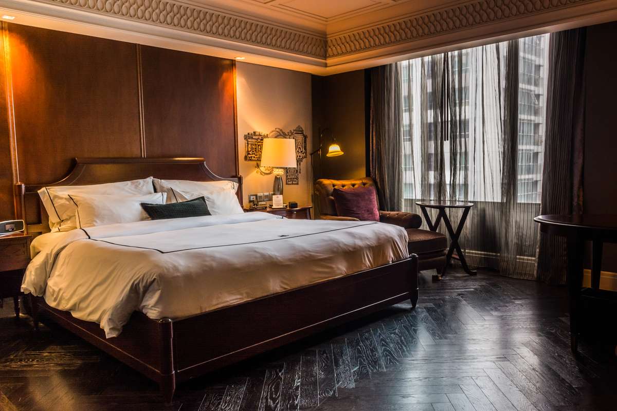 A hotel room with a large bed and white linens, warm wall sconces, dark wood floors, and a window with sheer curtains.