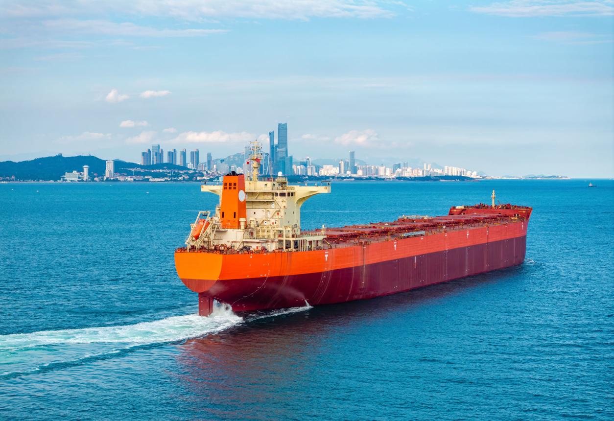 A bulk carrier under way at sea with a city skyline in the distance.