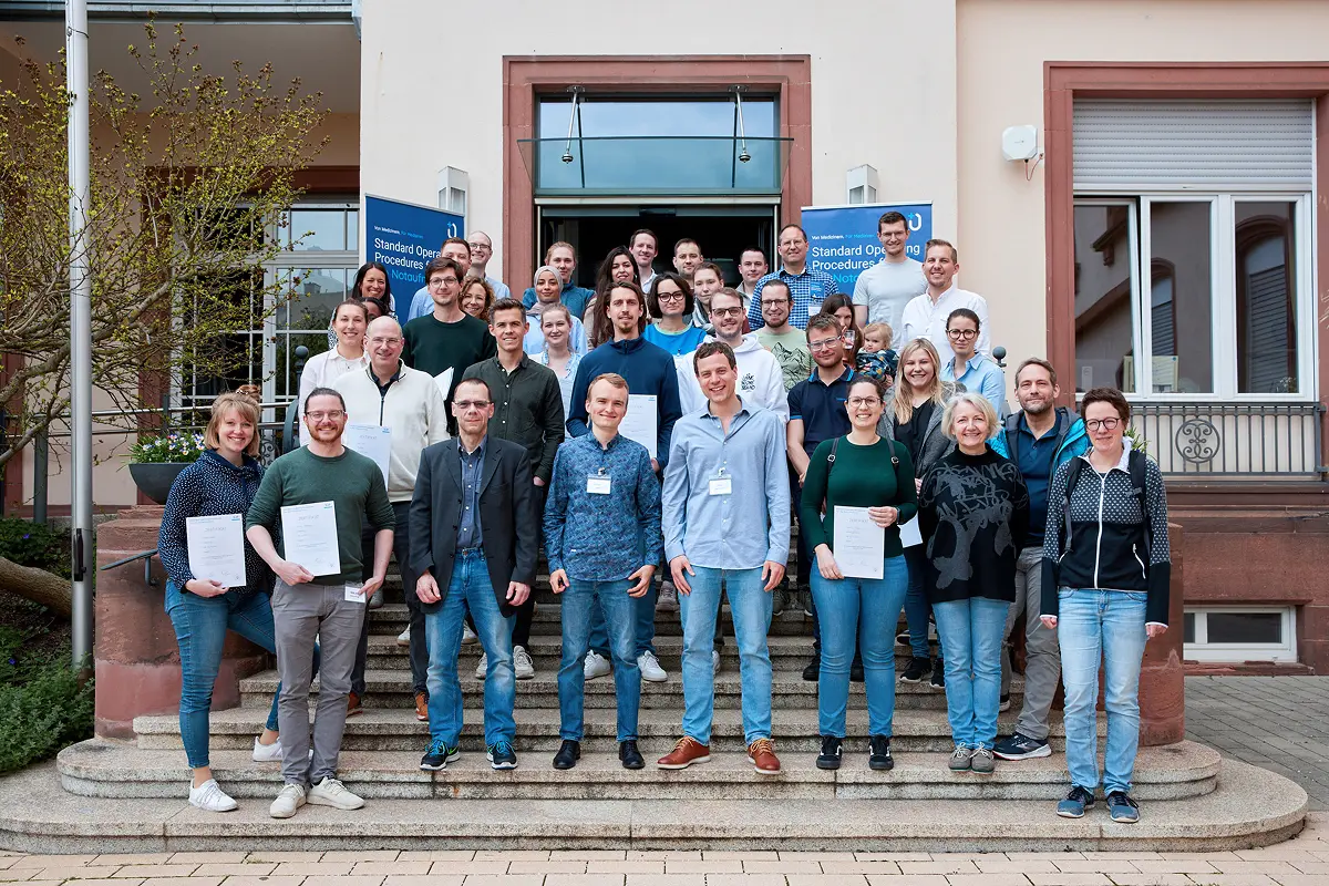 Absolventen des SOP Notaufnahme Seminars 2023 stehen auf den Stufen eines Gebäudes. Einige halten Zertifikate in der Hand, vor ihnen hängen Bildungs-Banner. 