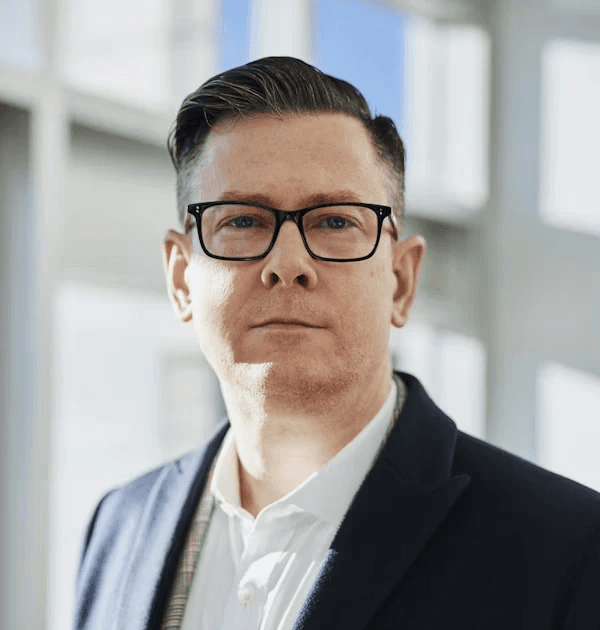 Professional headshot of a man with glasses and a dark blazer in a bright, blurred office setting.