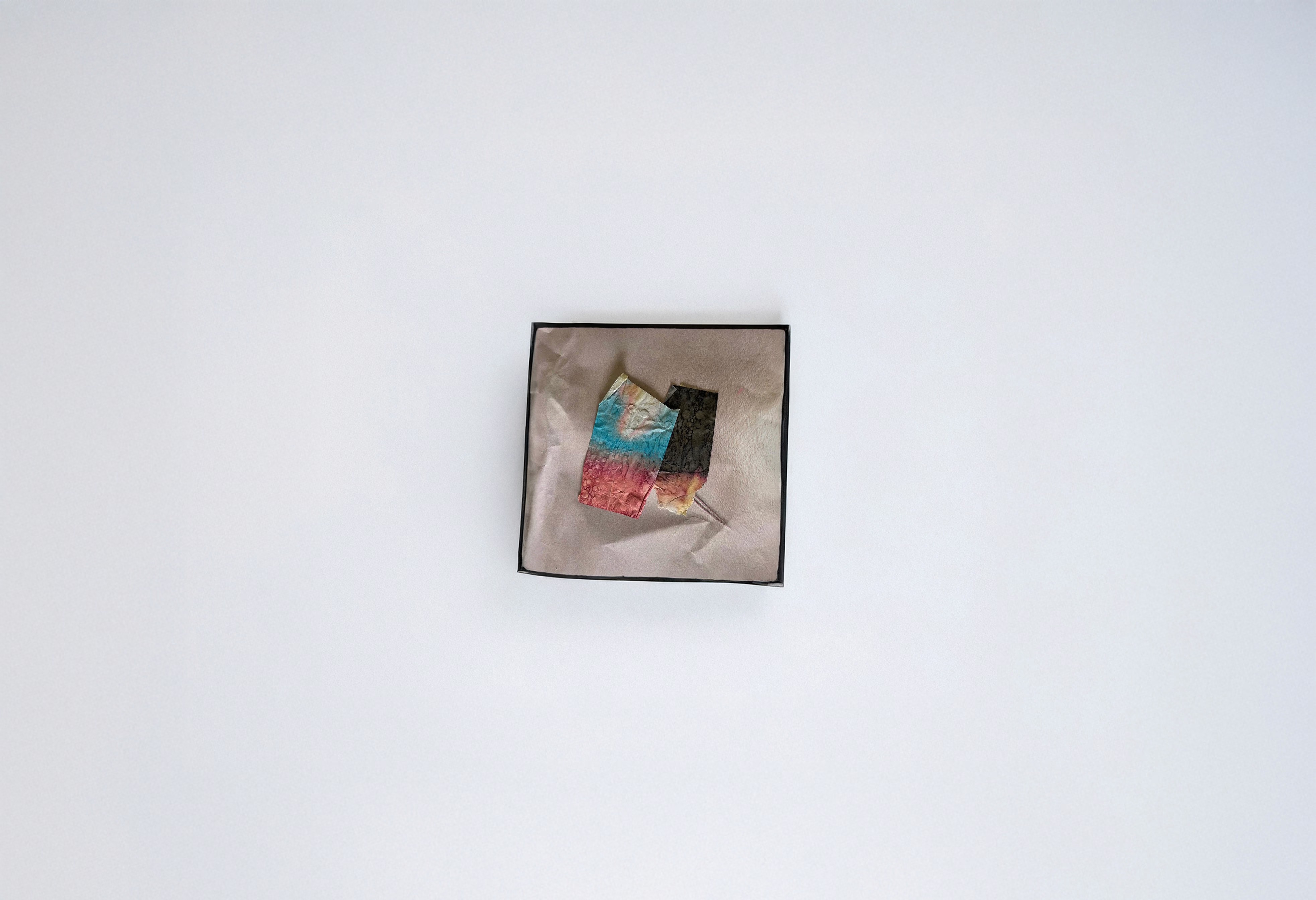 A small box holding two textured paper fragments—one dyed with gradients of blue, pink, and gold, the other dark and marbled—carefully placed on soft fabric. The composition feels intimate and archival, as if preserving remnants of memory, emotion, and tactile history.