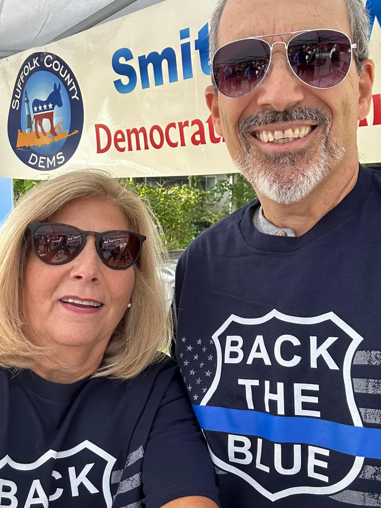 A man and a woman wearing sunglasses and matching shirts that say "BACK THE BLUE" pose together outdoors.