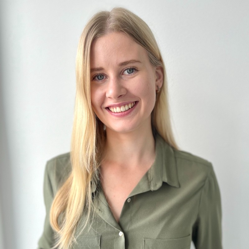 A young man with light brown hair is wearing a black suit jacket and white shirt, standing in a softly lit office setting, conveying professionalism.