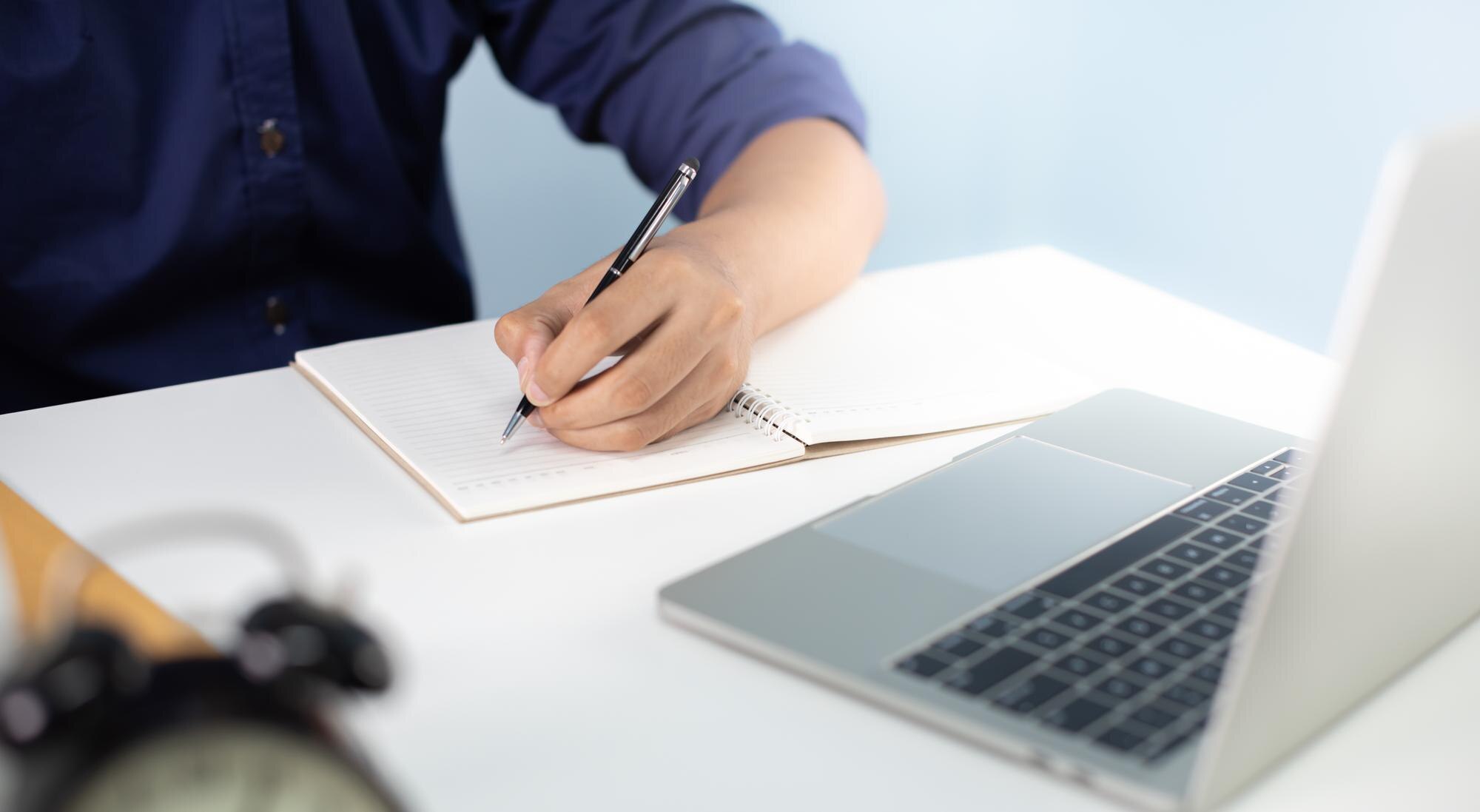 Close-up of a person writing in a lined notebook at a desk, with an open laptop beside the notebook and a blurred alarm clock in the foreground.