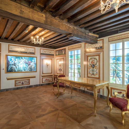 Elegáns szoba díszes fa mennyezettel, csillárokkal, nagy ablakokkal, aranyszegélyű asztallal, vörös bársony székekkel és faliképekkel.
