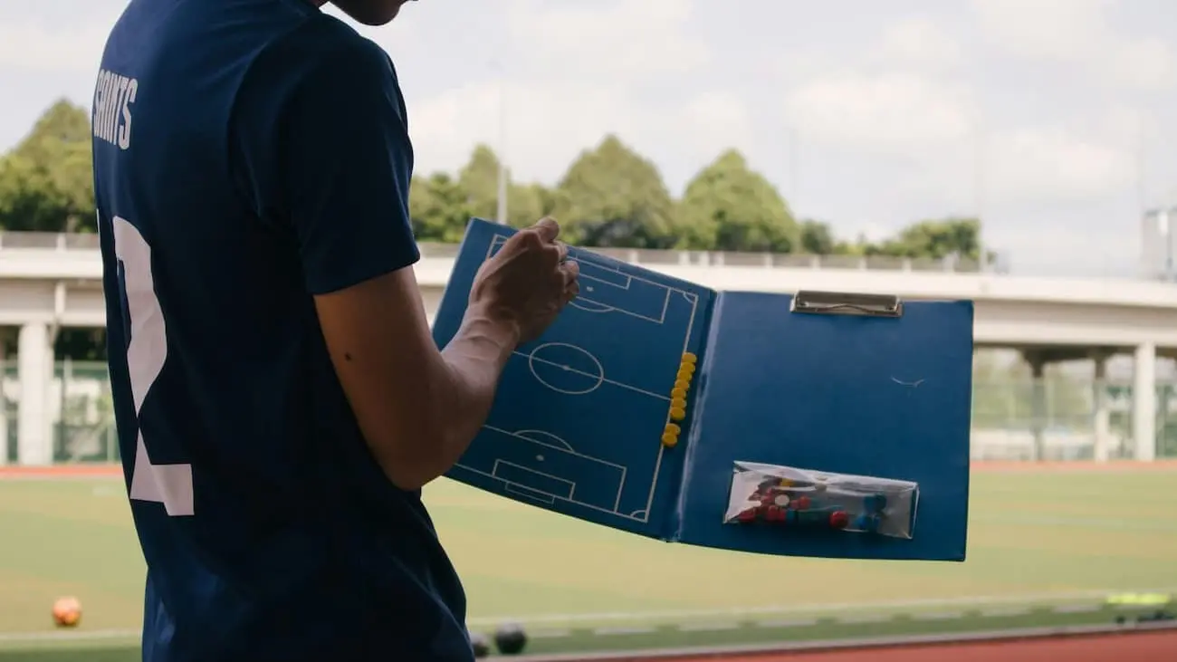 A football coach in a dark blue T-shirt holding a tactics board stands on a football pitch. The coach is planning the game strategy during training, an example of the practical challenges in coach training following the reforms of the German Football Association (DFB).