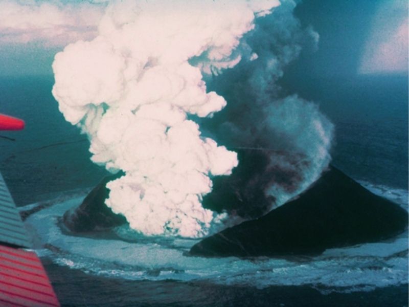  An overhead view of Surtsey, a volcanic eruption on a new island in the sea off the coast of Iceland. Ash and steam pour out of the newly formed volcanic crater and basalt begins to form an island.