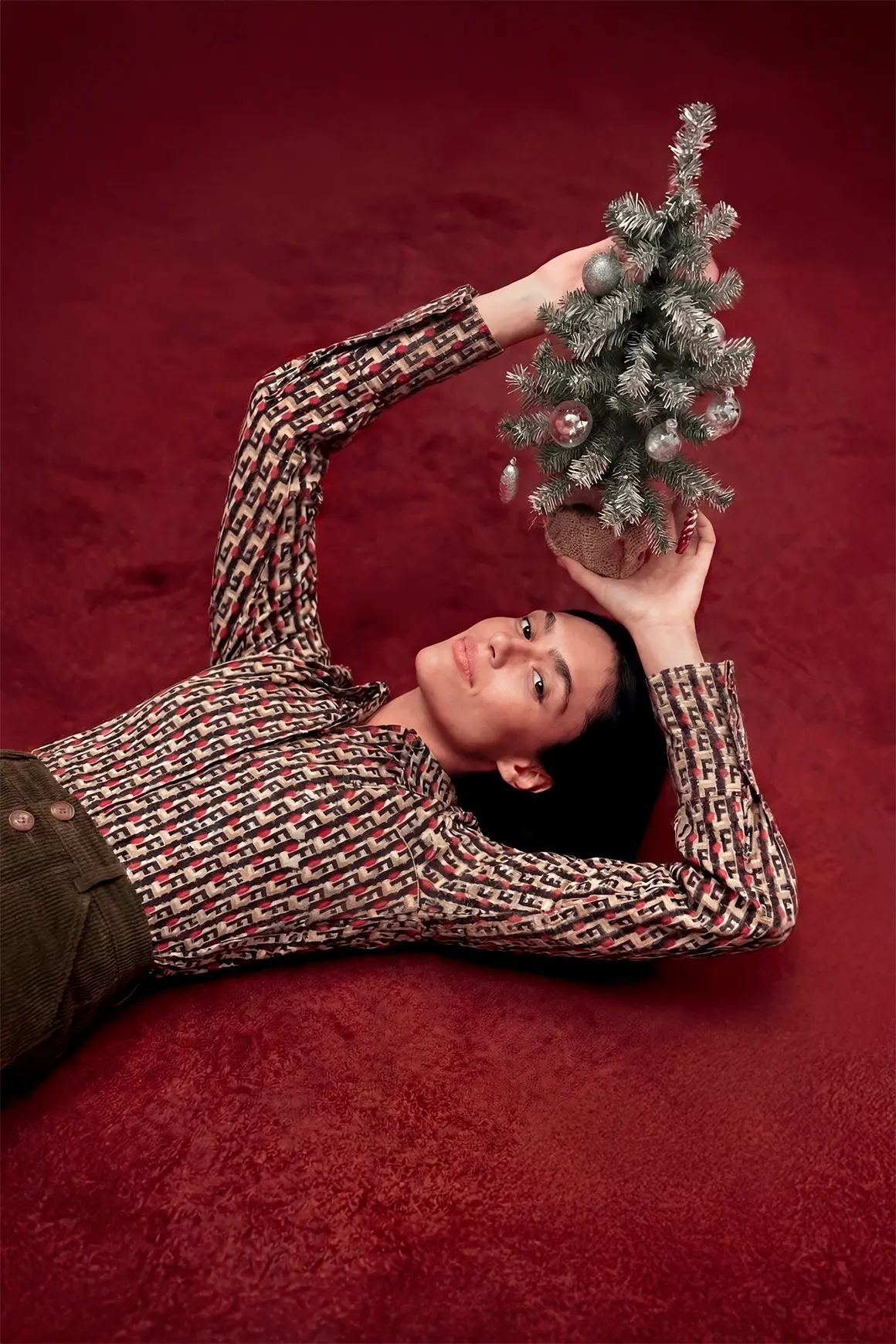 A person lies on a plush red carpet, holding a small decorated Christmas tree, wearing a patterned blouse and brown trousers, creating a luxurious scene ideal for a high-end fashion brand's AI production project.