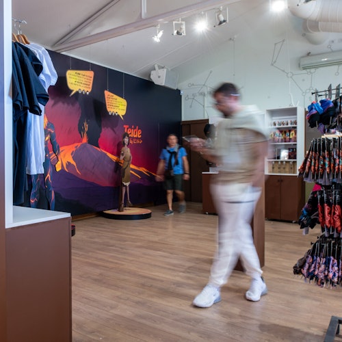An interior of a store with racks of clothing and souvenirs, a mural on the wall, and two blurred people walking by.
