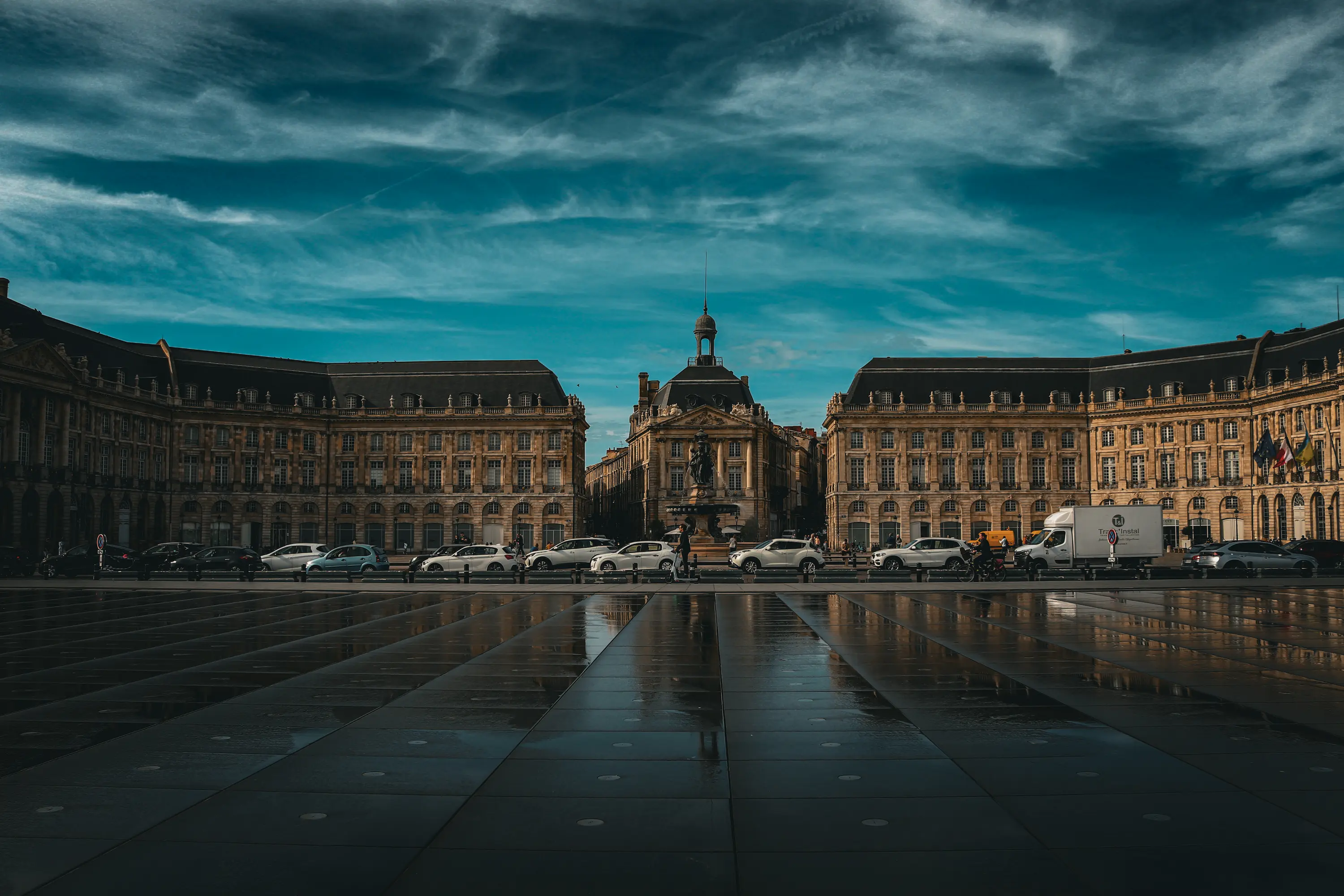 Vue de la Place de la Bourse à Bordeaux sous un ciel nuageux, avec le Miroir d’eau au premier plan reflétant les façades
