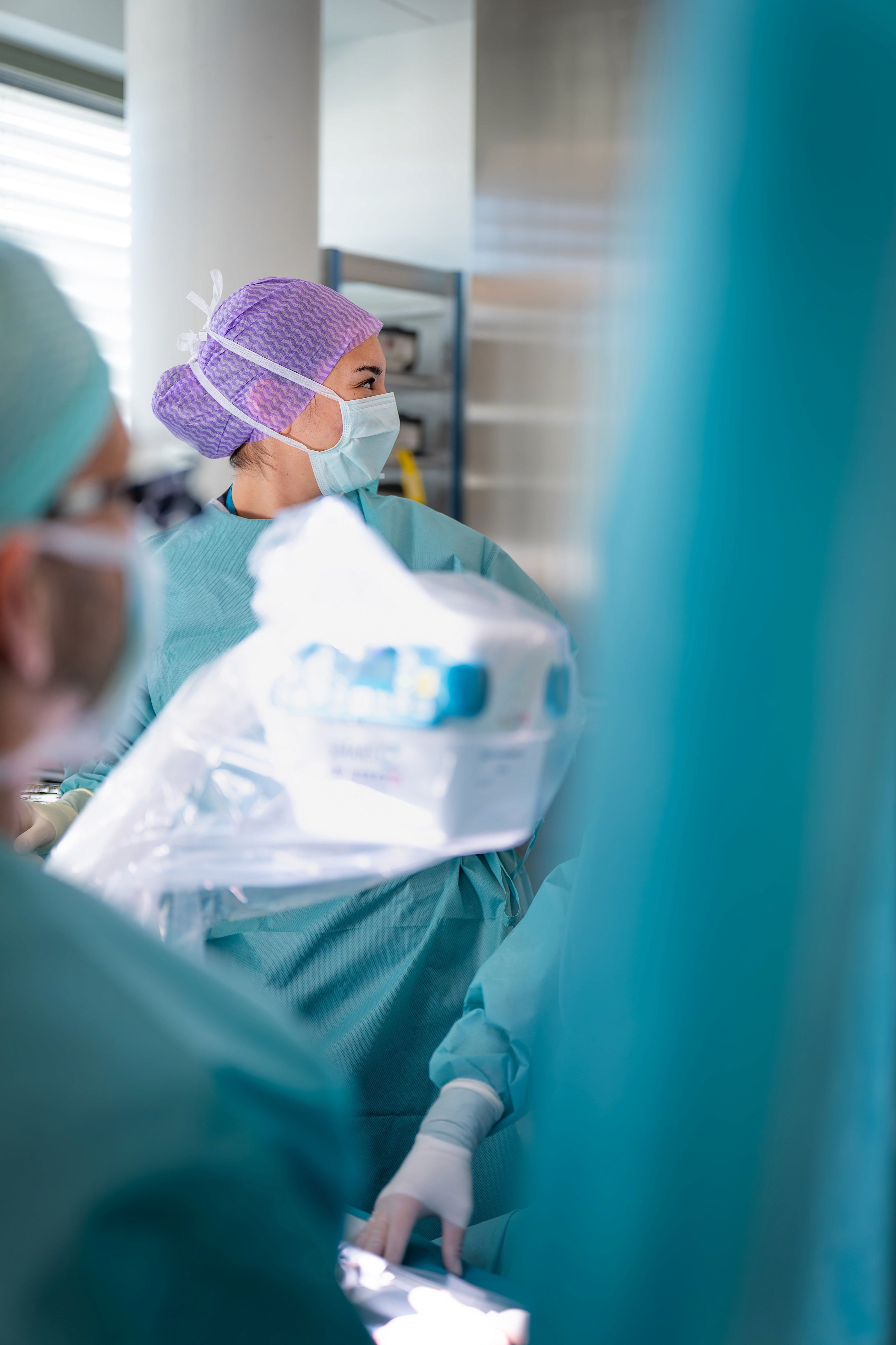 Eine medizinische Fachkraft mit Kittel, Maske und violettem Haarnetz sitzt in einem hell erleuchteten Spitalzimmer im Handzentrum Pratteln und hält Geräte in der Hand. Andere maskierte Fachkräfte sind teilweise im Vordergrund zu sehen.