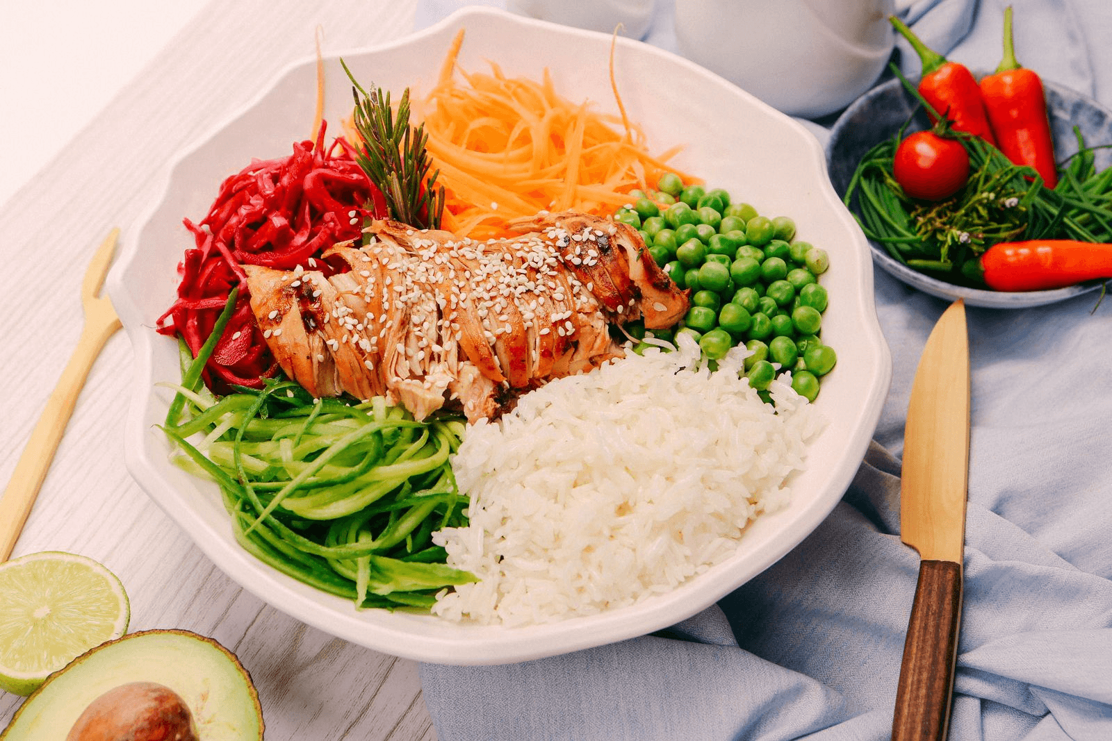 A colorful salad bowl with grilled chicken topped with sesame seeds, surrounded by white rice, shredded carrots, green peas, cucumber strips, and beetroot. The setting includes an avocado half, sliced lime, and a small plate of red chilies and herbs. A knife and fork are placed beside the plate on a light blue cloth, conveying a fresh and vibrant meal.