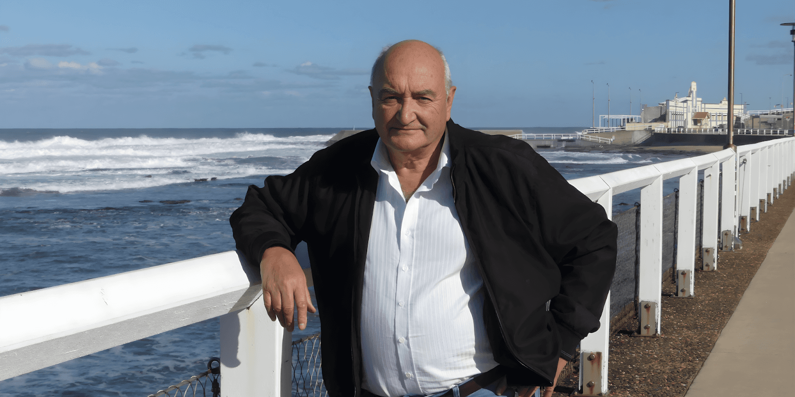 A man stands on a pier by the ocean, wearing a black jacket, white shirt, and jeans, looking relaxed.