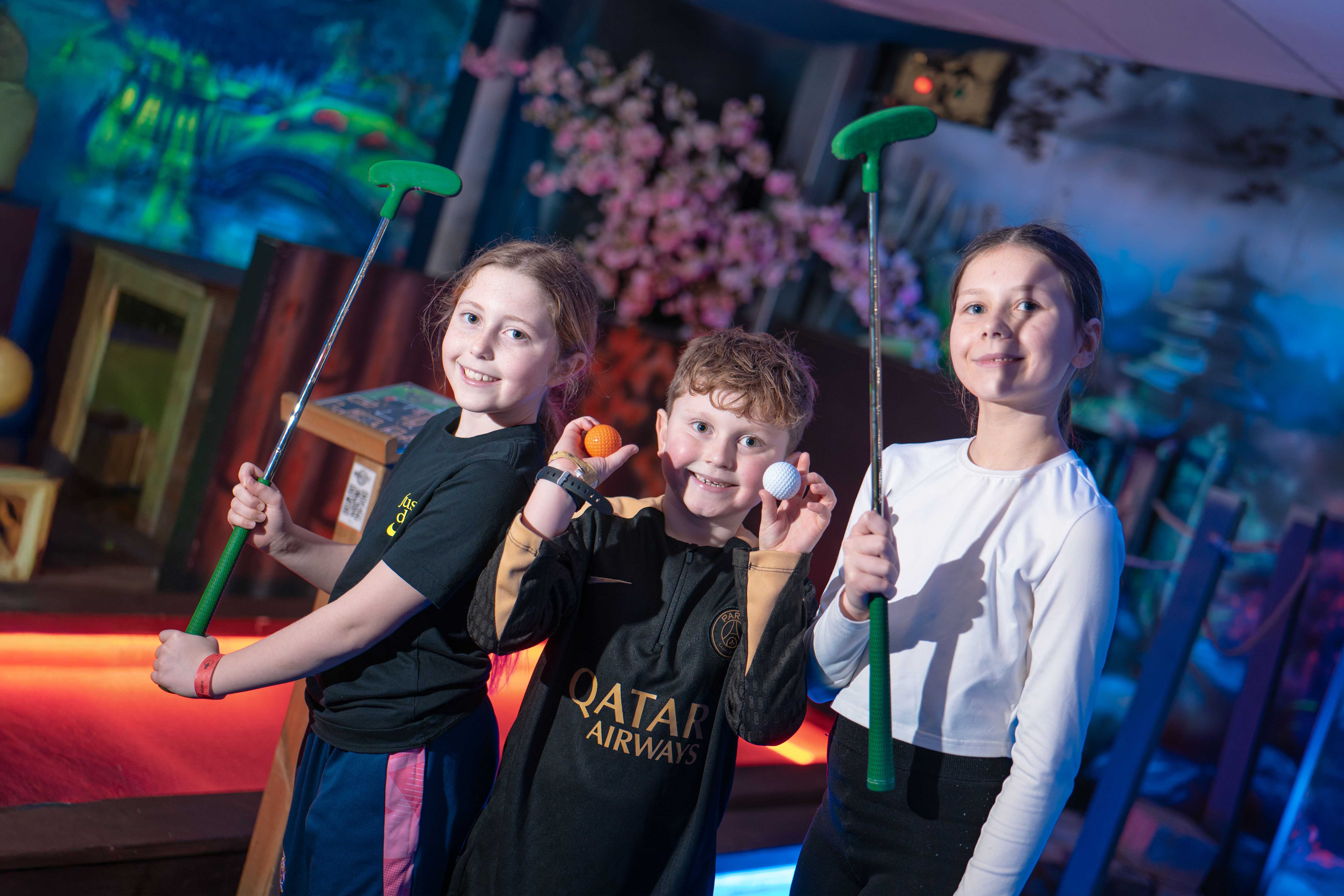 Three children smile for the camera while holding golf putters and balls at an indoor mini golf course.
