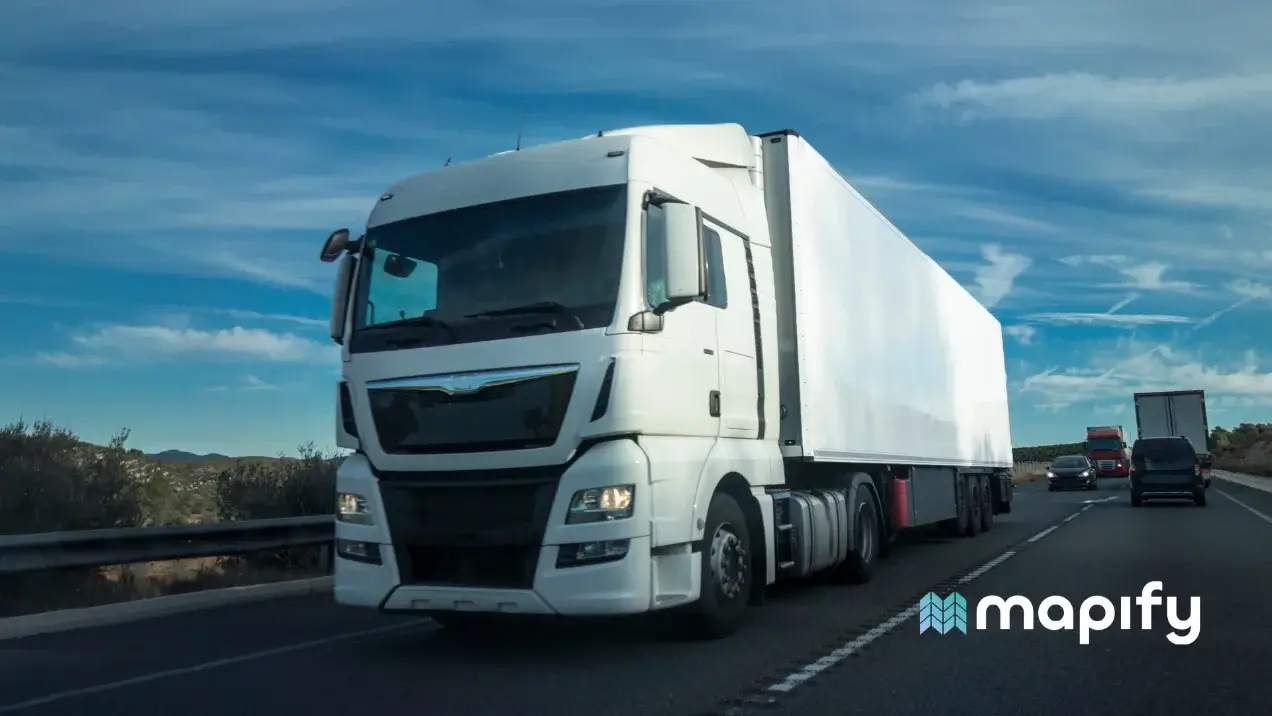 A large white semi-truck travels down a highway under a clear blue sky. Other vehicles follow behind. The road is bordered by low vegetation.