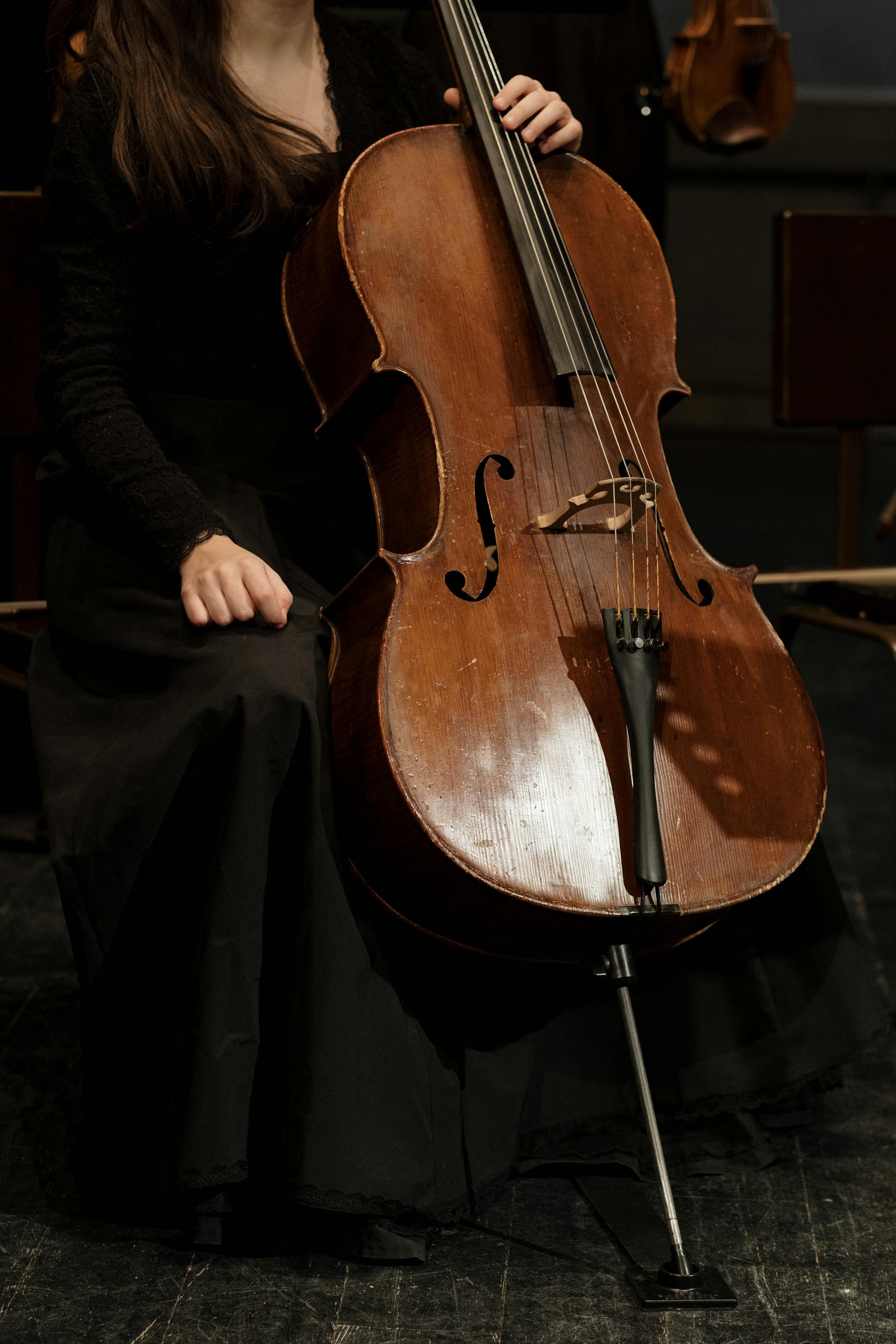 A close-up of a musician in a black lace-sleeved dress and long skirt seated with a wooden cello resting between her knees.