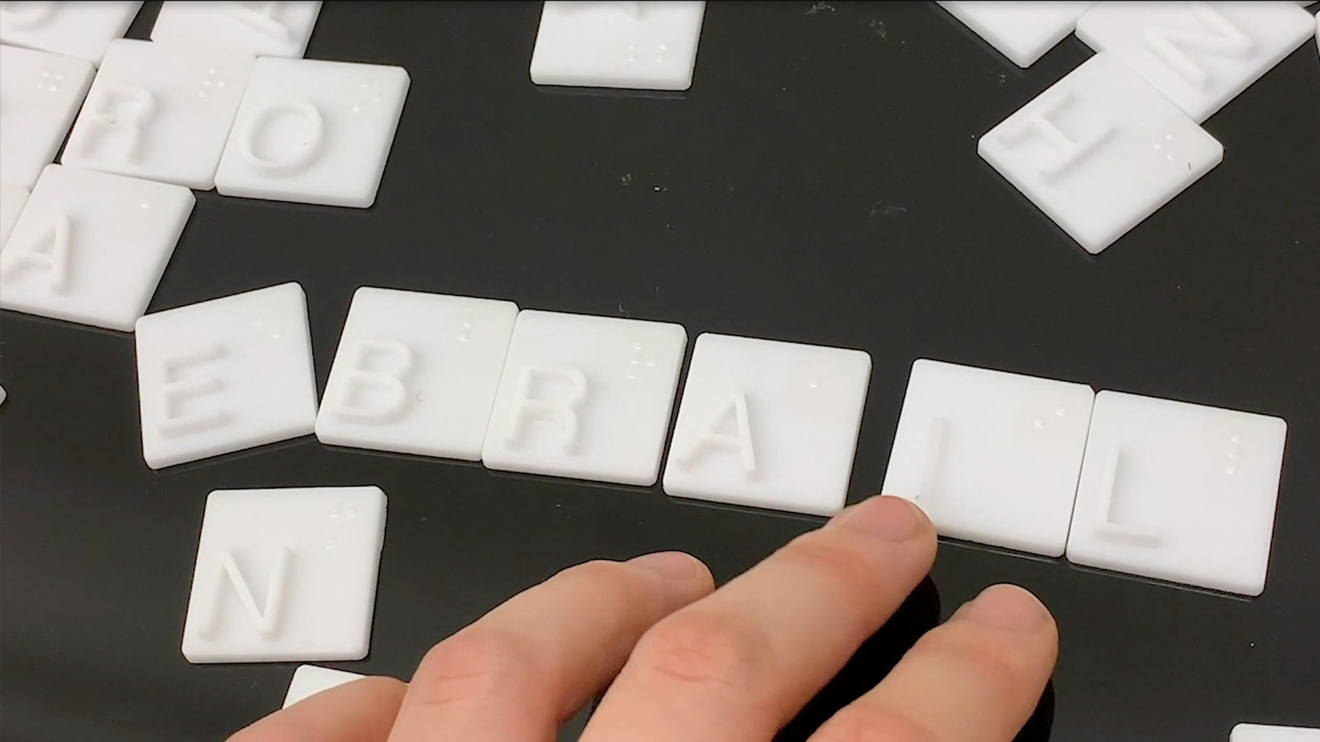 A hand is positioned over a black surface covered with white letter tiles arranged in rows. The tiles spell out “BAL”, “E”, “N”, “O”, “R”, and “T”. The hand’s index finger rests on the “L” tile.