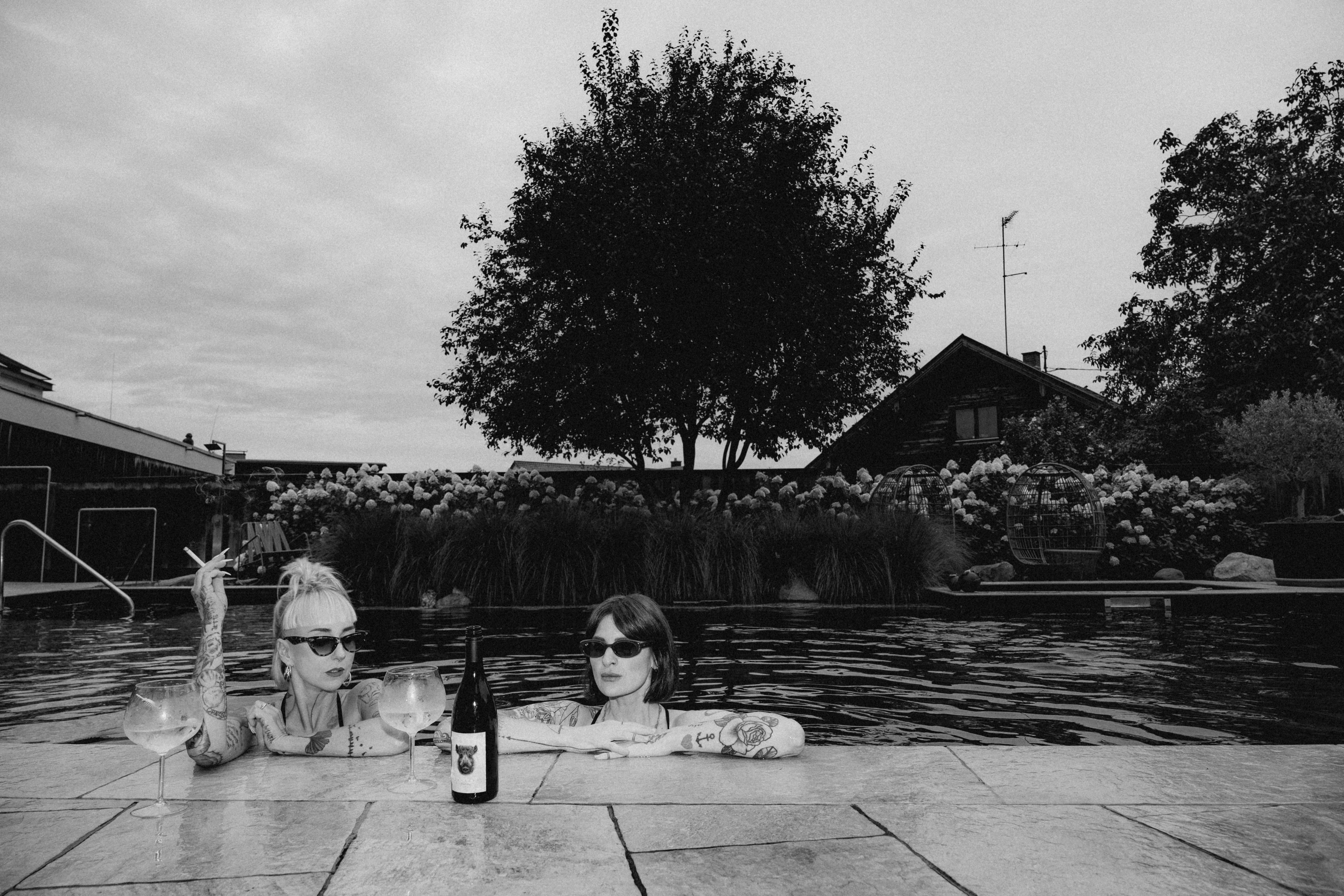 Two people walk by a body of water with a tree and a house in the background, captured in black and white.