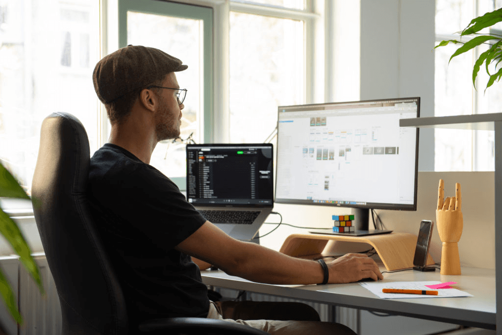 Mann mit Brille und Schiebermütze sitzt an einem modernen Schreibtisch in einem hellen Büro. Er arbeitet an einem großen Monitor, während ein Laptop daneben geöffnet ist.