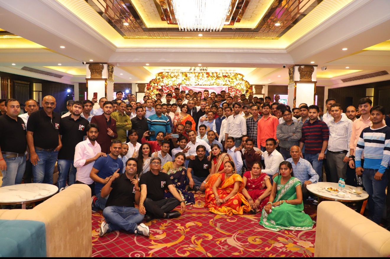 A young person with curly hair smiles at the camera while sitting in front of a laptop against a dark background.