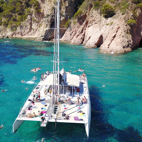 Un catamaran ancré dans une eau bleu clair près de falaises rocheuses, avec des personnes nageant et prenant le soleil sur le bateau.