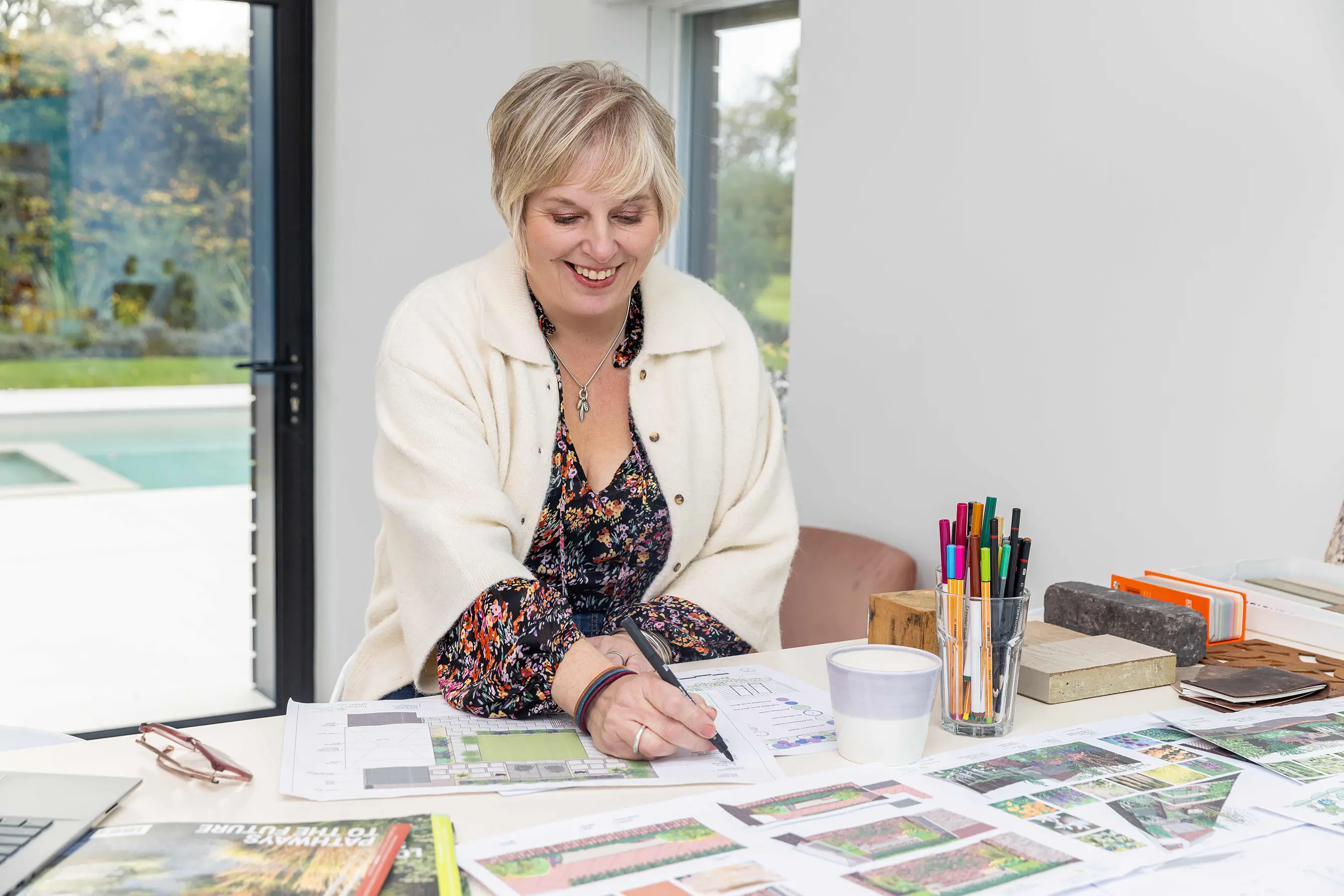 A woman with short hair sits at a table covered in papers and a coffee cup, smiling in a bright, modern room.