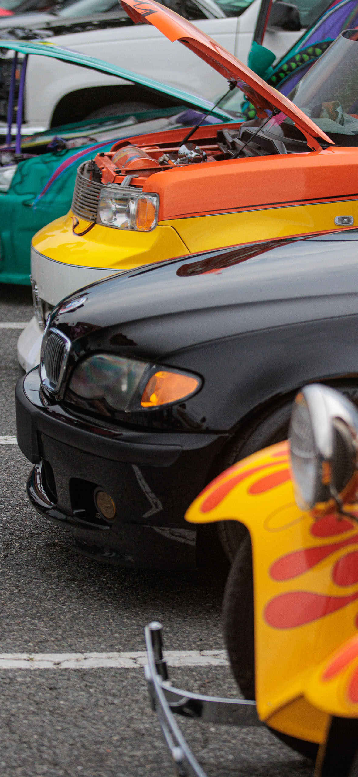 Close-up of several cars parked in a row, including a black BMW, yellow and orange custom cars, and a teal car.