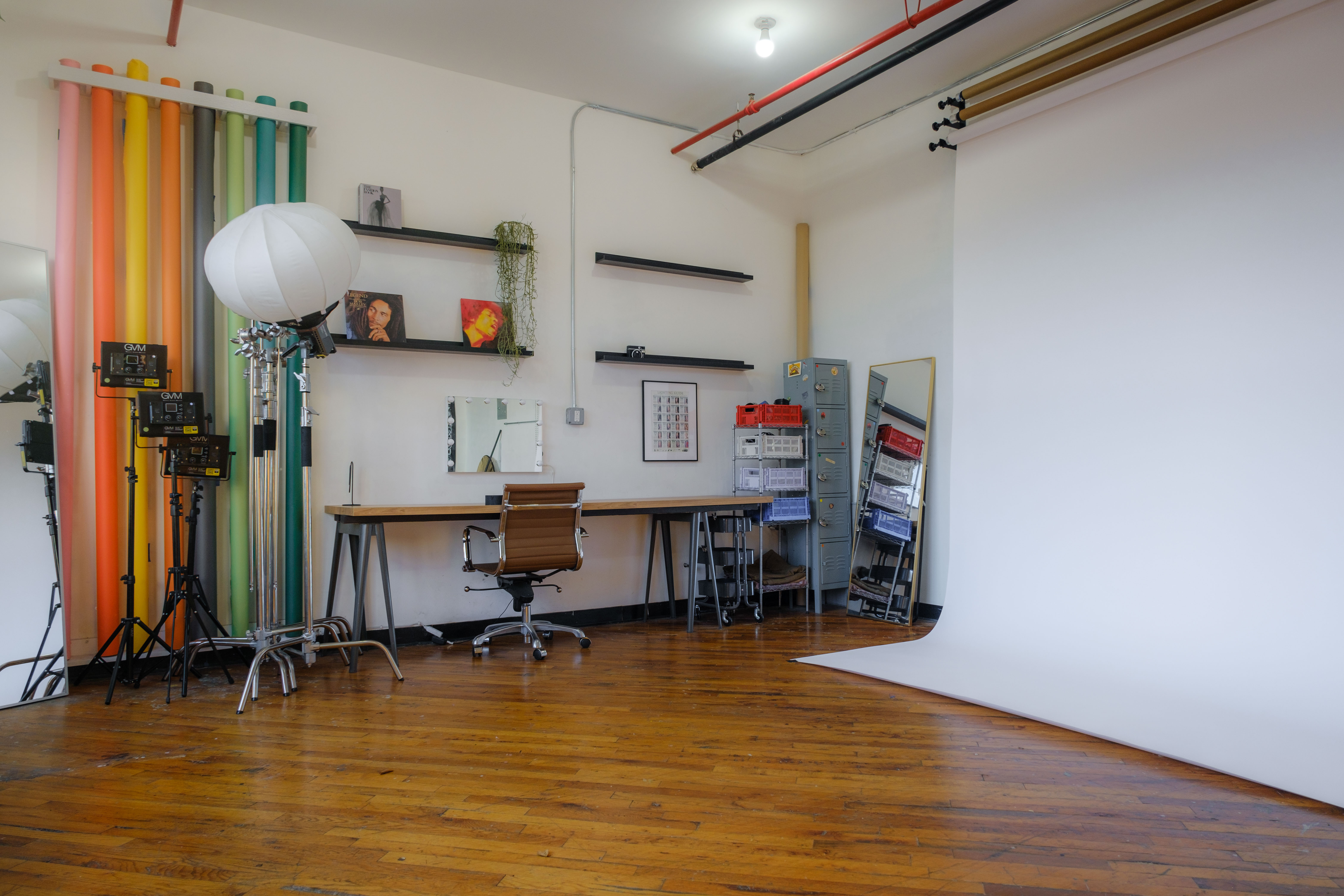 A daylight photography studio featuring a large wall of colorful backdrops, professional lighting equipment, and a workspace with shelves displaying decor and camera gear. A floral wall installation and a large mirror add artistic elements to the studio’s bright, versatile environment.