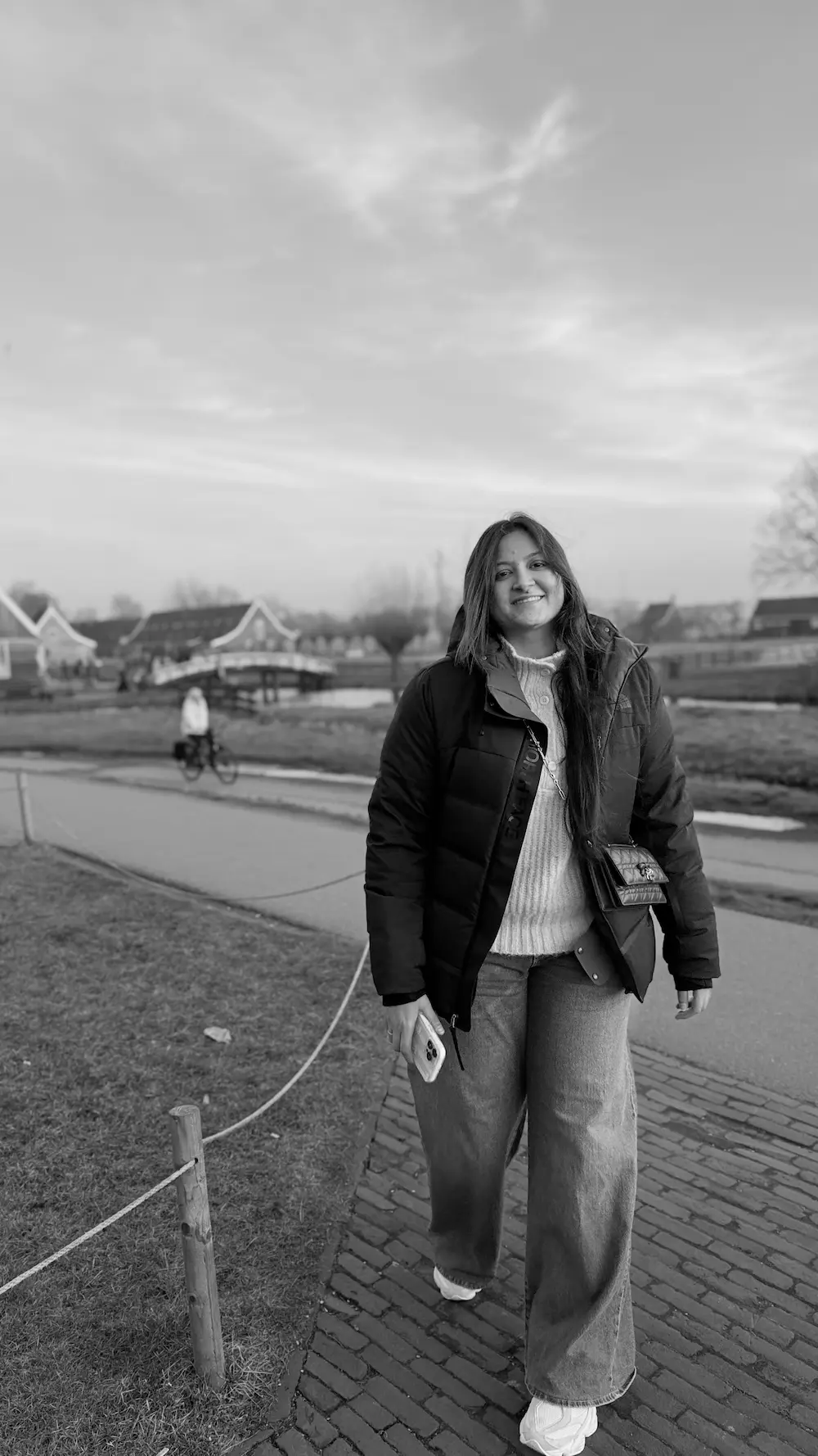 A person in a winter jacket and wide-leg pants walks along a brick path in a serene neighborhood, with traditional houses and a canal softly illuminated by the setting sun in the background.