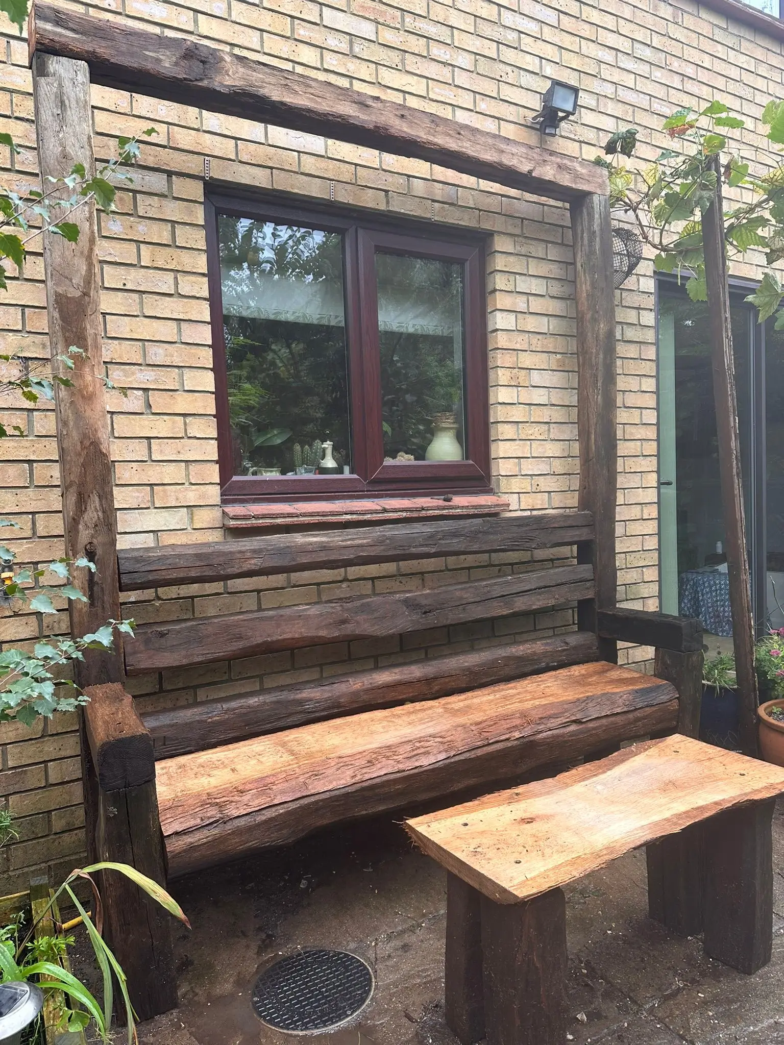 A wooden bench and table set against a brick wall with a window, surrounded by plants.