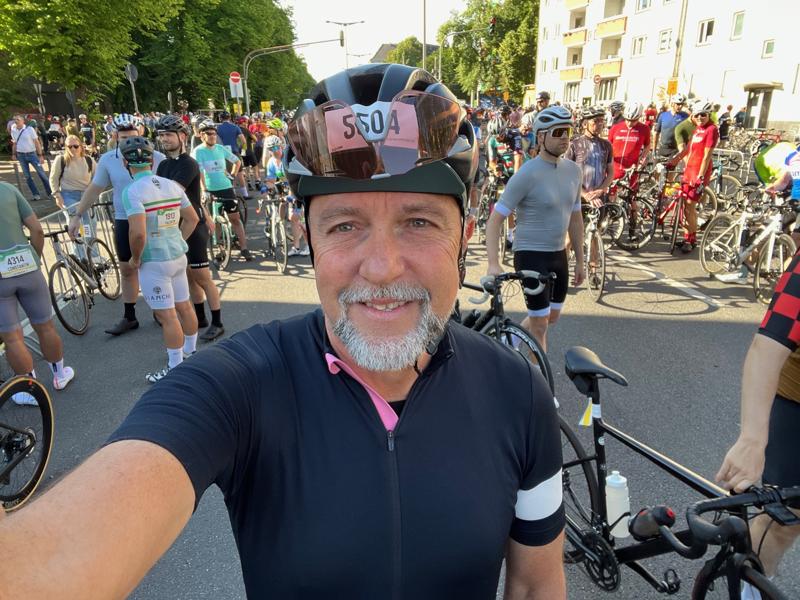 A cyclist wearing a black helmet and jersey takes a selfie in front of a large group of cyclists gathered with their bicycles on a sunny day, ready for a race or event.