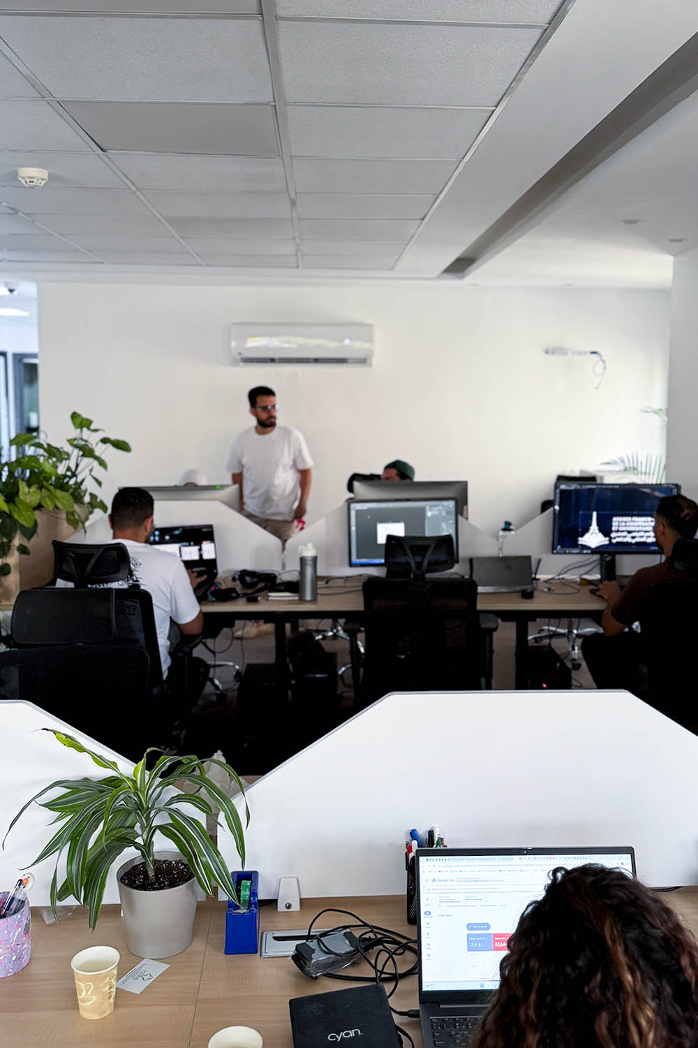 A presenter stands in front of an audience in an office setting, with laptops open on the tables.
