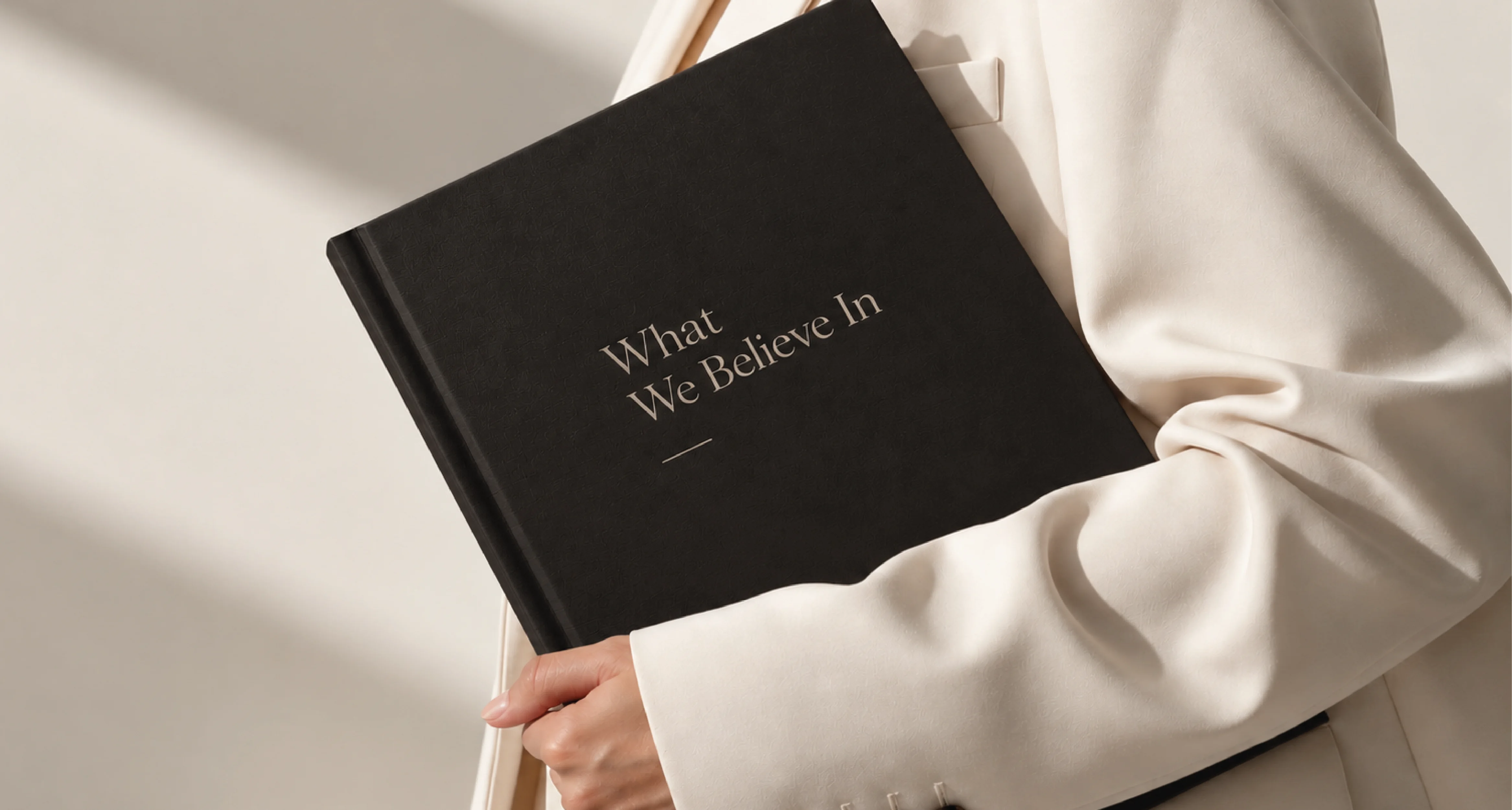 A person in a white suit holds a black book titled "What We Believe In," capturing a sense of elegance and professionalism.