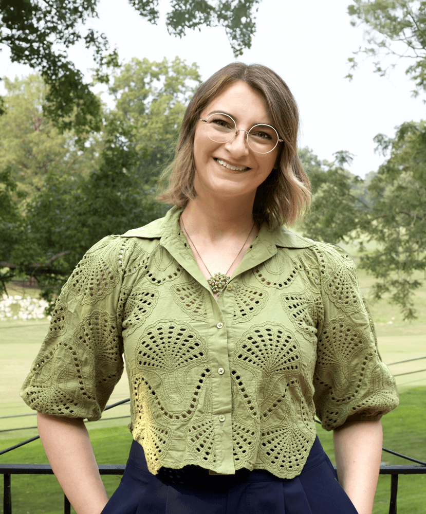 A smiling woman with shoulder-length blonde hair and round glasses sits outdoors wearing a sage green eyelet lace blouse with intricate cutout patterns and three-quarter sleeves, paired with a navy skirt and delicate necklace. She's positioned against a natural backdrop of lush green trees and grass on what appears to be a sunny day.