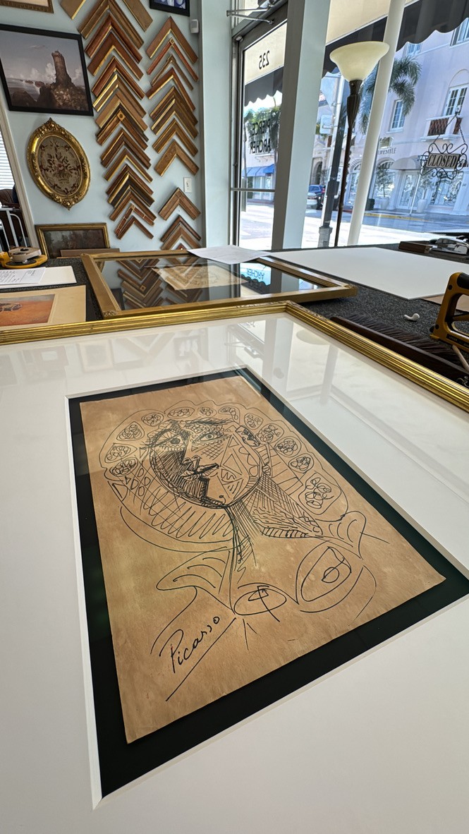 Picasso-signed line drawing of a face laid on a table for framing, with frame corner samples on the wall behind